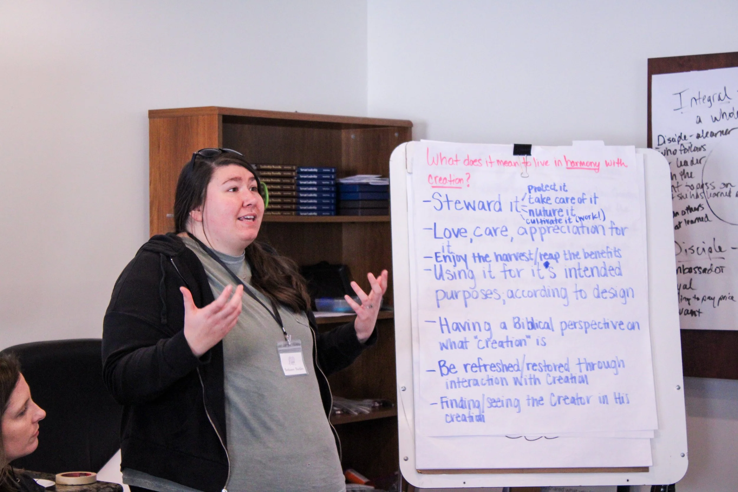 A woman with long brown hair and glasses on her head is speaking in front of a whiteboard filled with handwritten notes. She's gesturing with her hands, wearing a black hoodie and a gray t-shirt. Other people are partially visible in the room.
