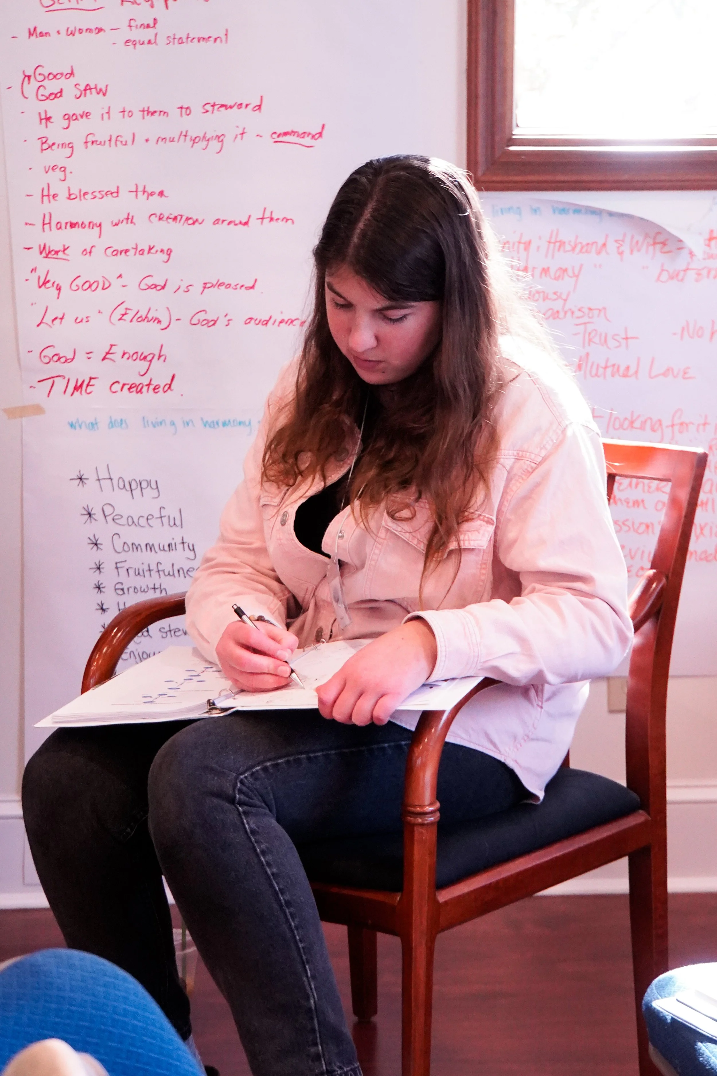 A young woman sitting in a wooden chair takes notes in a notebook during a class or workshop. She is wearing a light pink jacket and has long, wavy brown hair. Behind her, there are whiteboards filled with handwritten notes in red and blue ink, with 