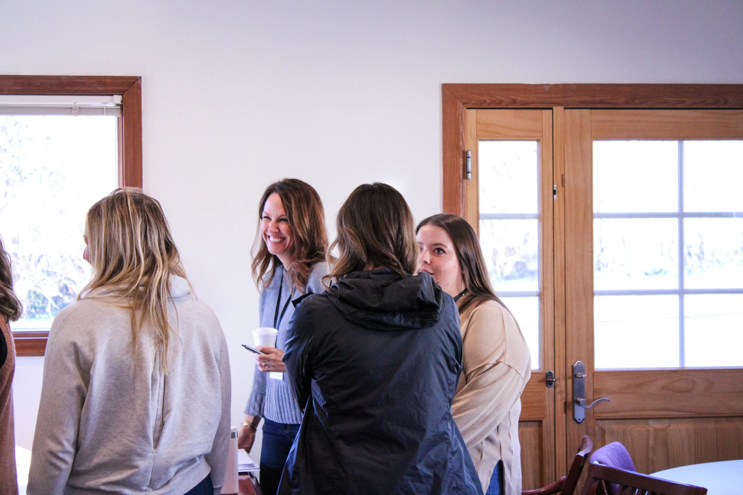 Group of women standing in a room with wooden framed windows and a wooden door, engaged in conversation. One woman is smiling and holding a cup and pen.