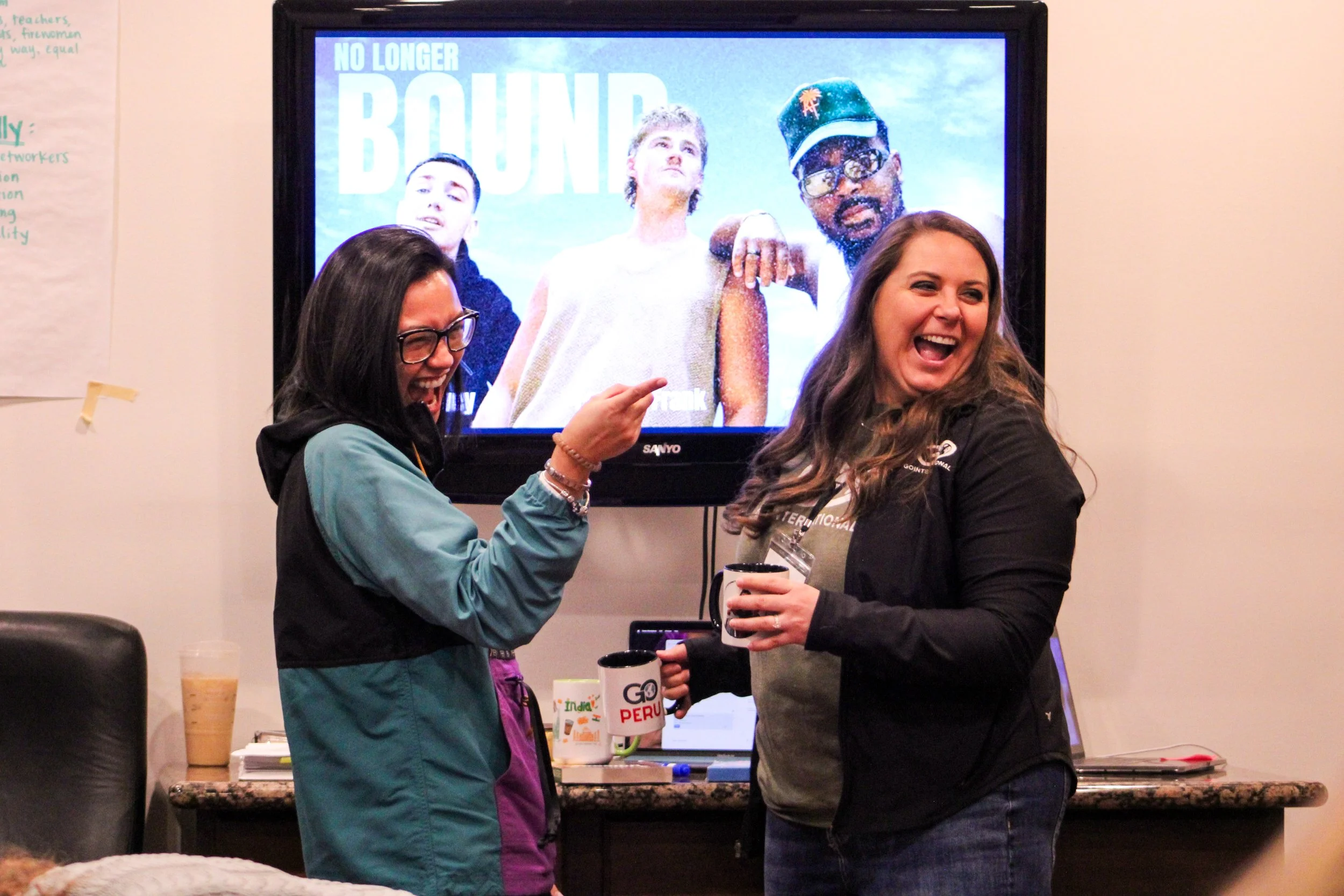 Two women laughing and talking in front of a television screen showing a promotional poster for a show titled 'No Longer BOUND' featuring three people posing with serious expressions. One woman is pointing at the poster.