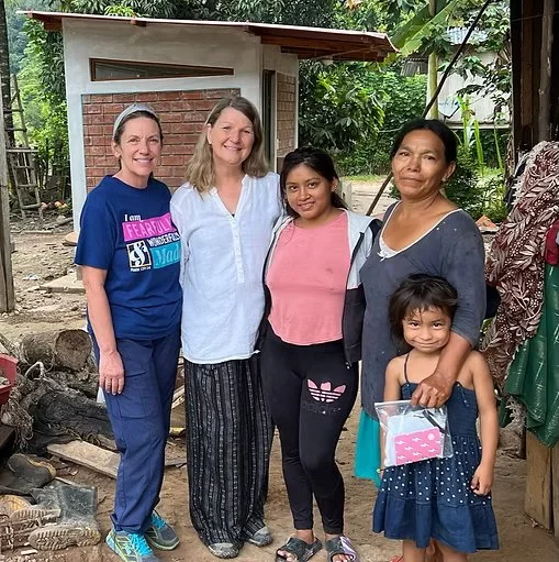Four women and a young girl standing outdoors in a rural area, smiling for the camera, with a small building and trees in the background.