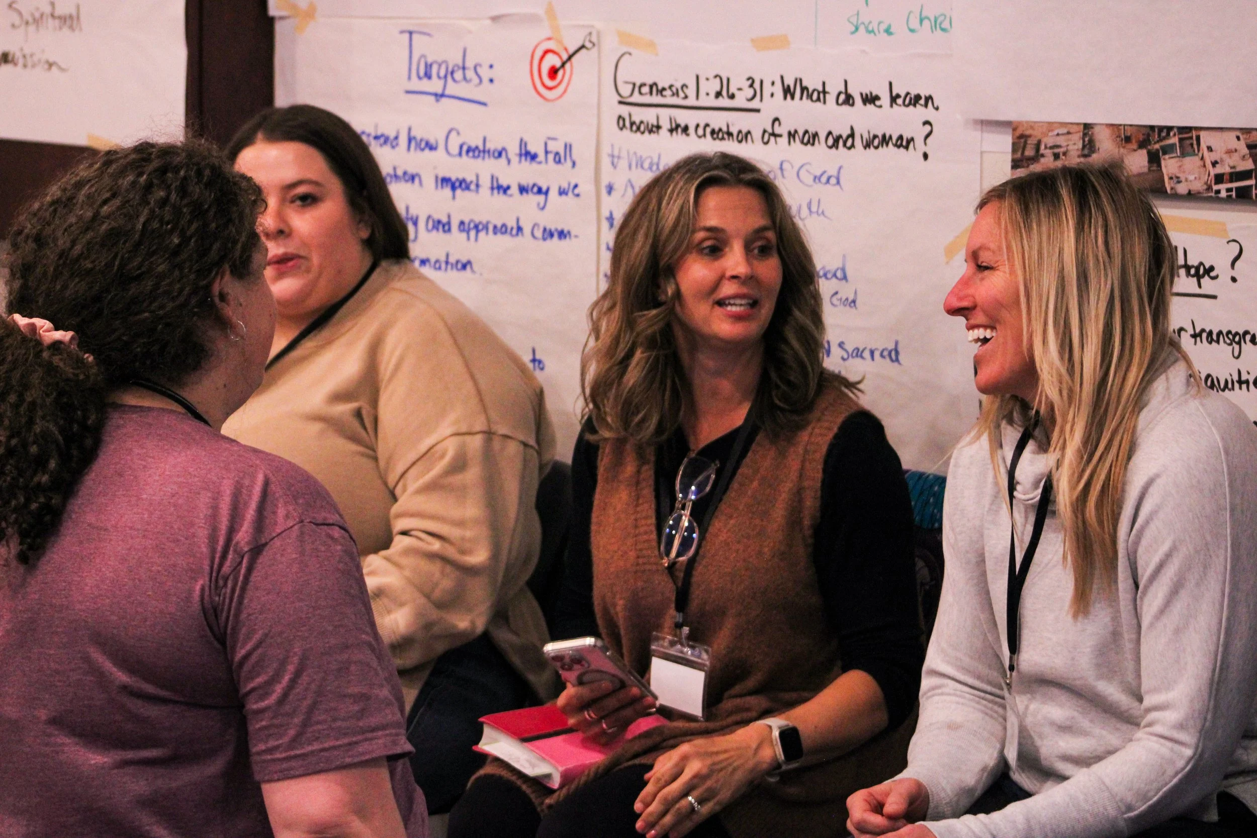 Four women are engaged in a conversation at an indoor event, with a large poster about biblical topics behind them.