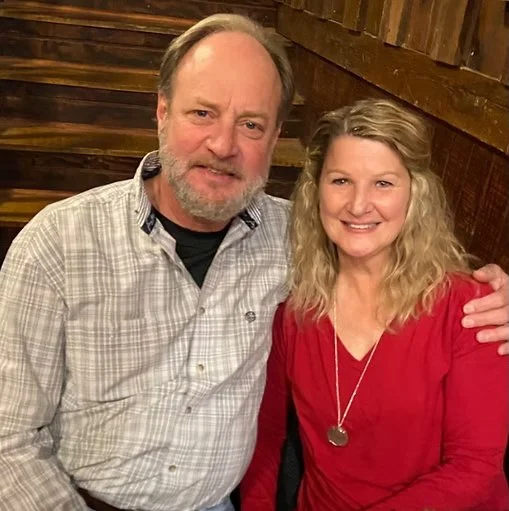 A man with a beard wearing a light plaid shirt and a woman with blonde curly hair in a red top pose together indoors with a wooden wall background.