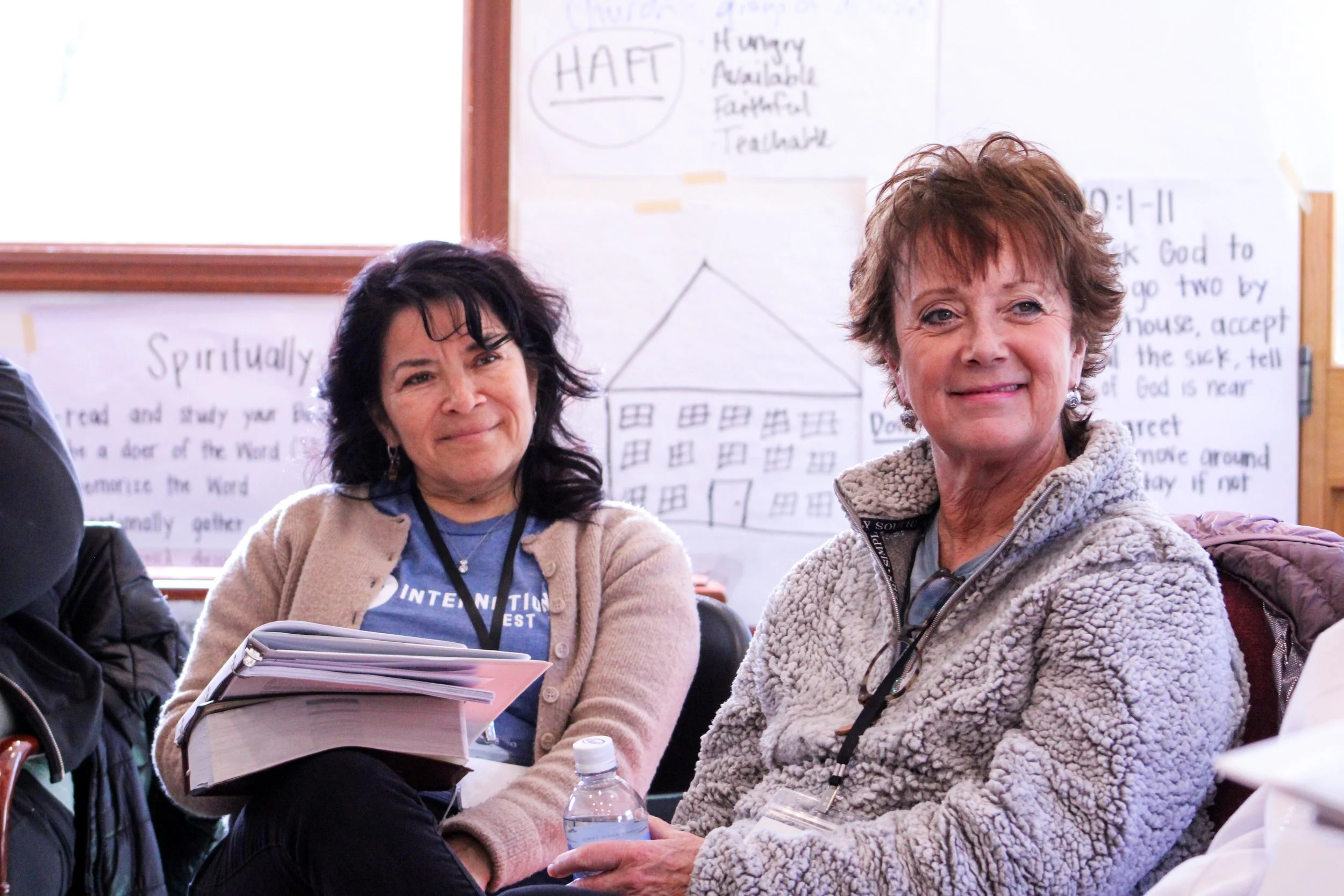 Two women sitting next to each other in a classroom or workshop setting, with educational posters and diagrams visible on the wall behind them.