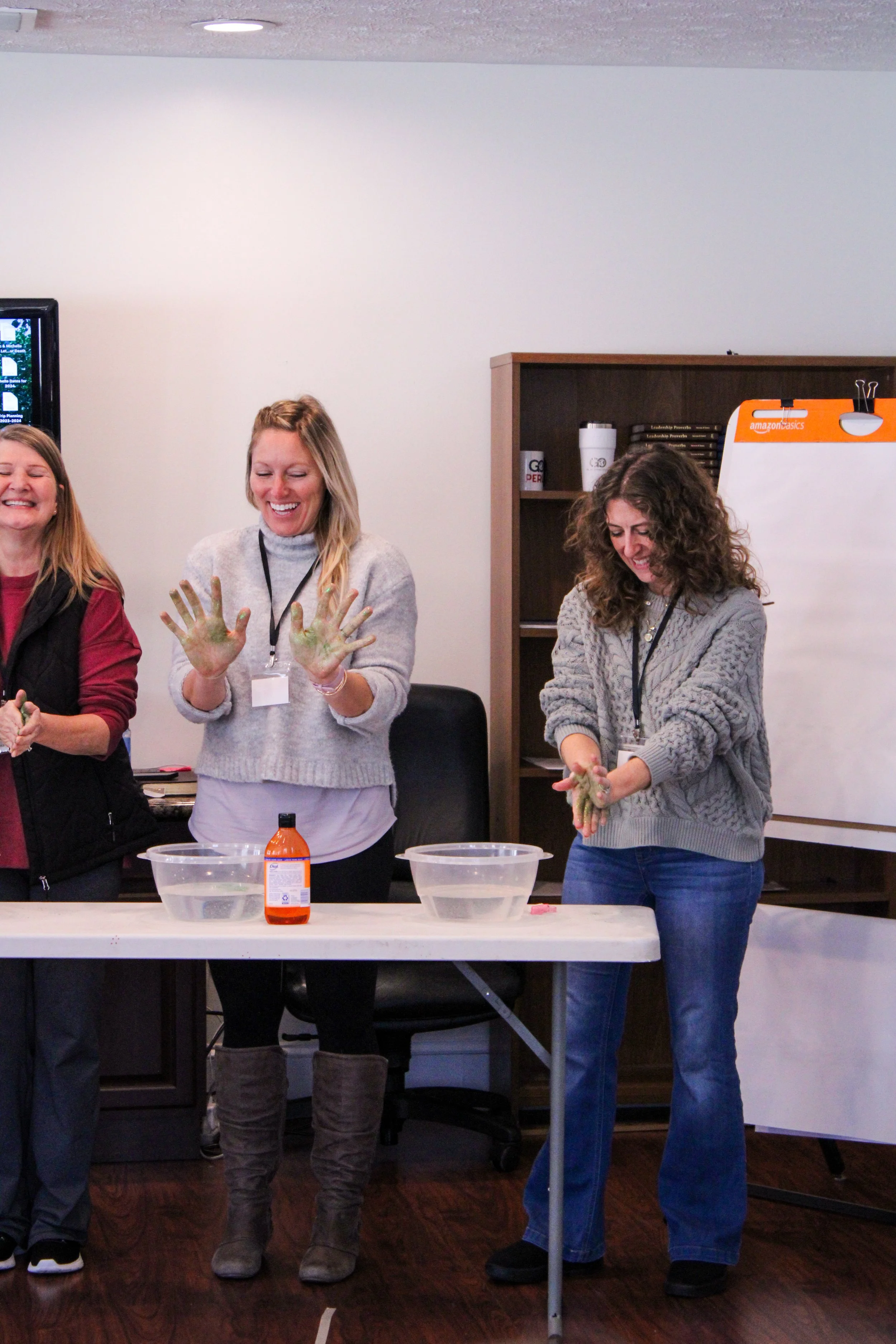 Three women standing at a table, two of them with their hands covered in a green substance, participating in a hands-on activity in a room. There are bowls and a bottle on the table, and a whiteboard in the background.