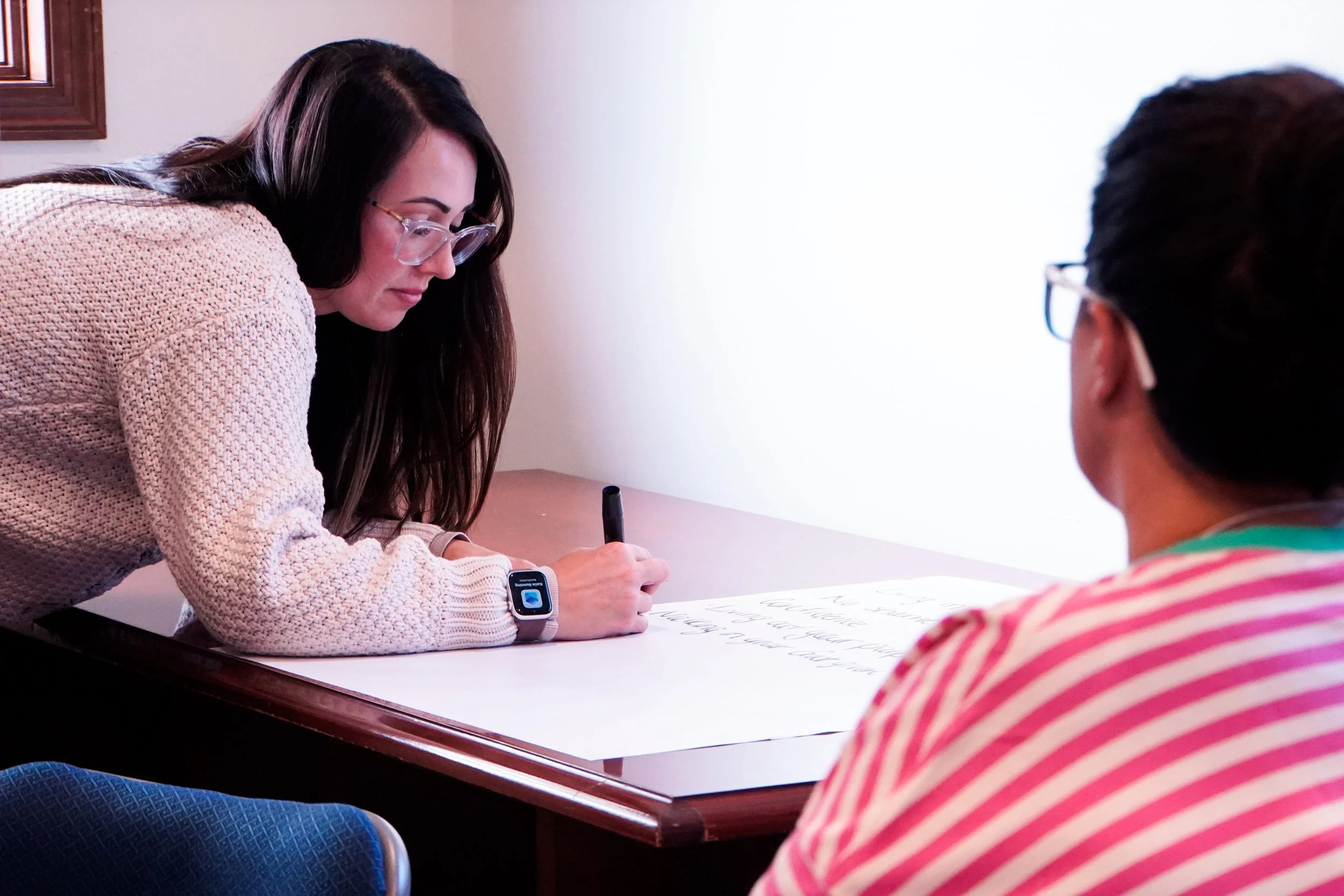 A woman with long dark hair, wearing glasses and a beige sweater, is sitting at a table with a notebook and a pen, writing. A person with short dark hair wearing glasses and a colorful striped shirt is sitting across from her. The setting appears to 