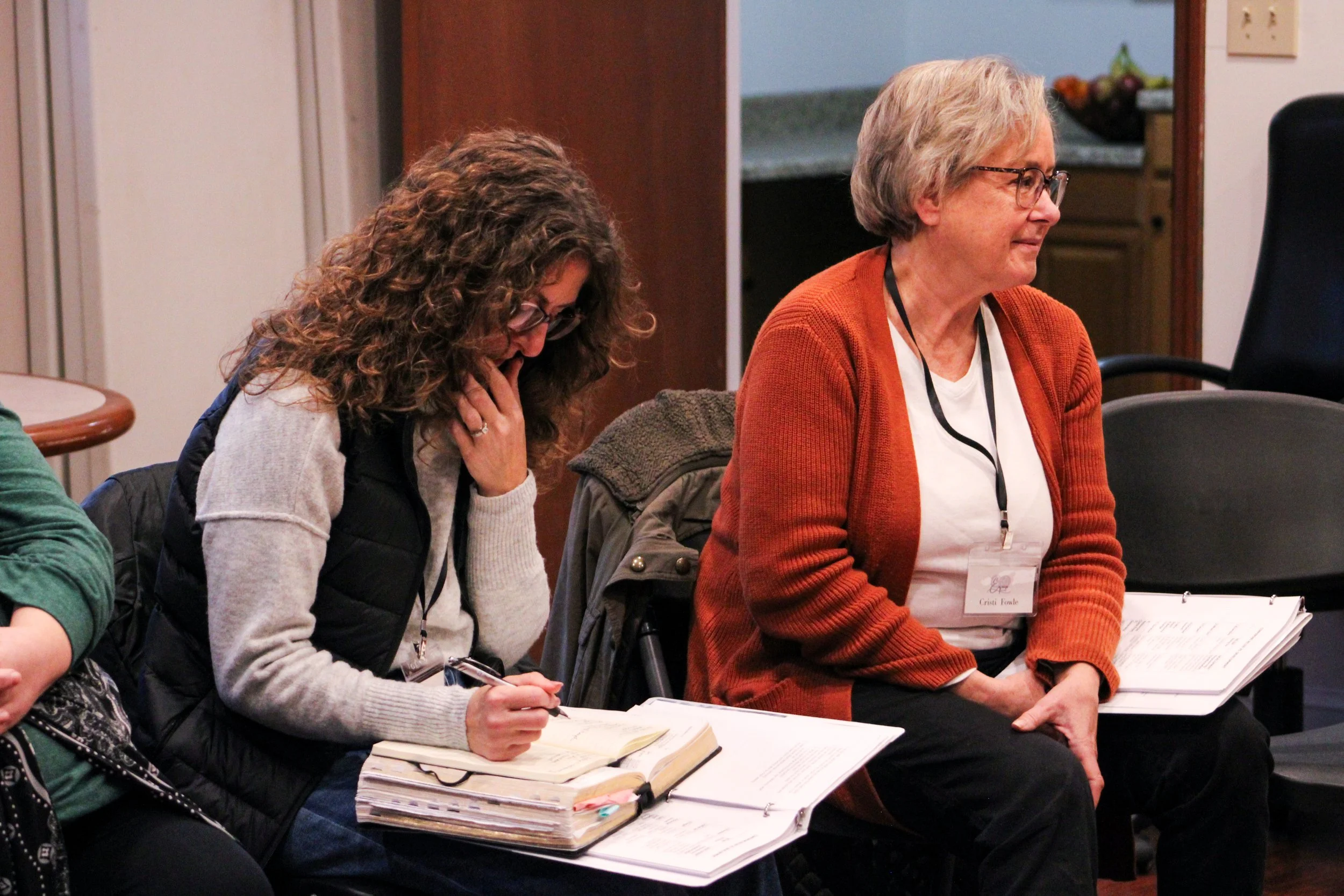 Two women seated at a table during a conference or workshop, one taking notes and the other listening, with papers and notebooks in front of them.