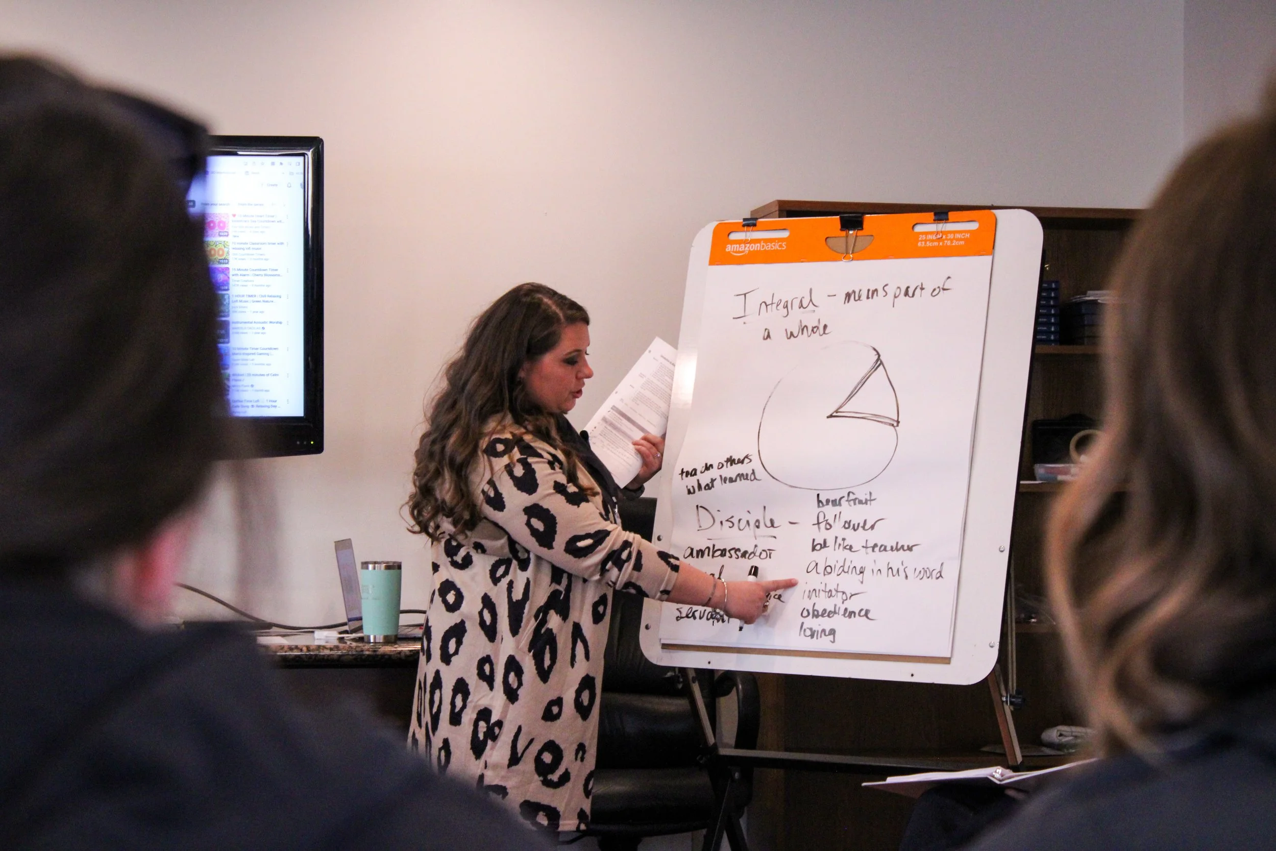 A woman with long, wavy brown hair leads a presentation in a room, standing next to a whiteboard with notes and a diagram about holistic concepts. Several people are watching her, with some heads visible in the foreground.