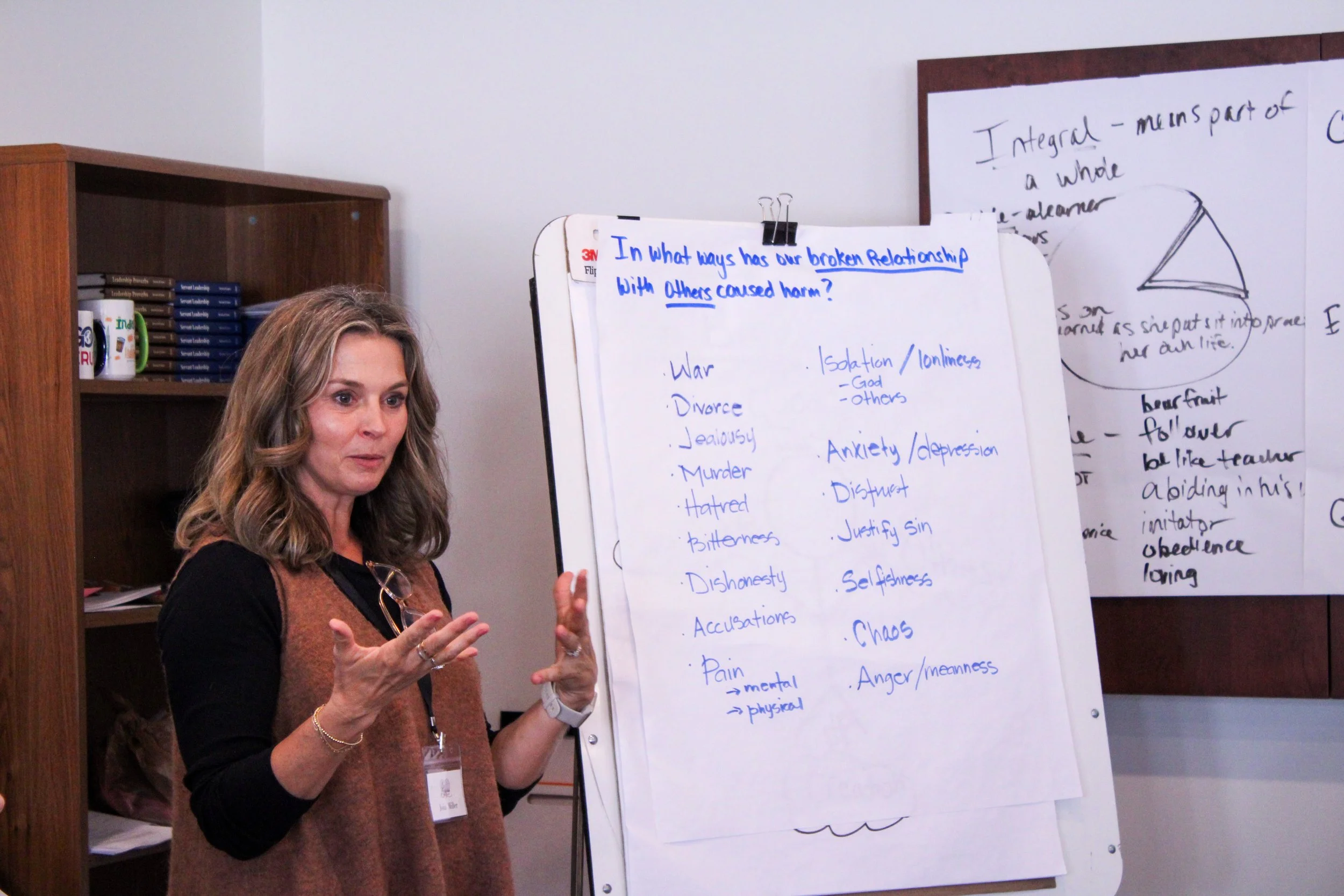 A woman with long, wavy blond hair, wearing a rust-colored vest over a black long-sleeve shirt, is standing in front of a whiteboard and flip chart while presenting. She appears to be explaining something, gesturing with her hands. The whiteboard has