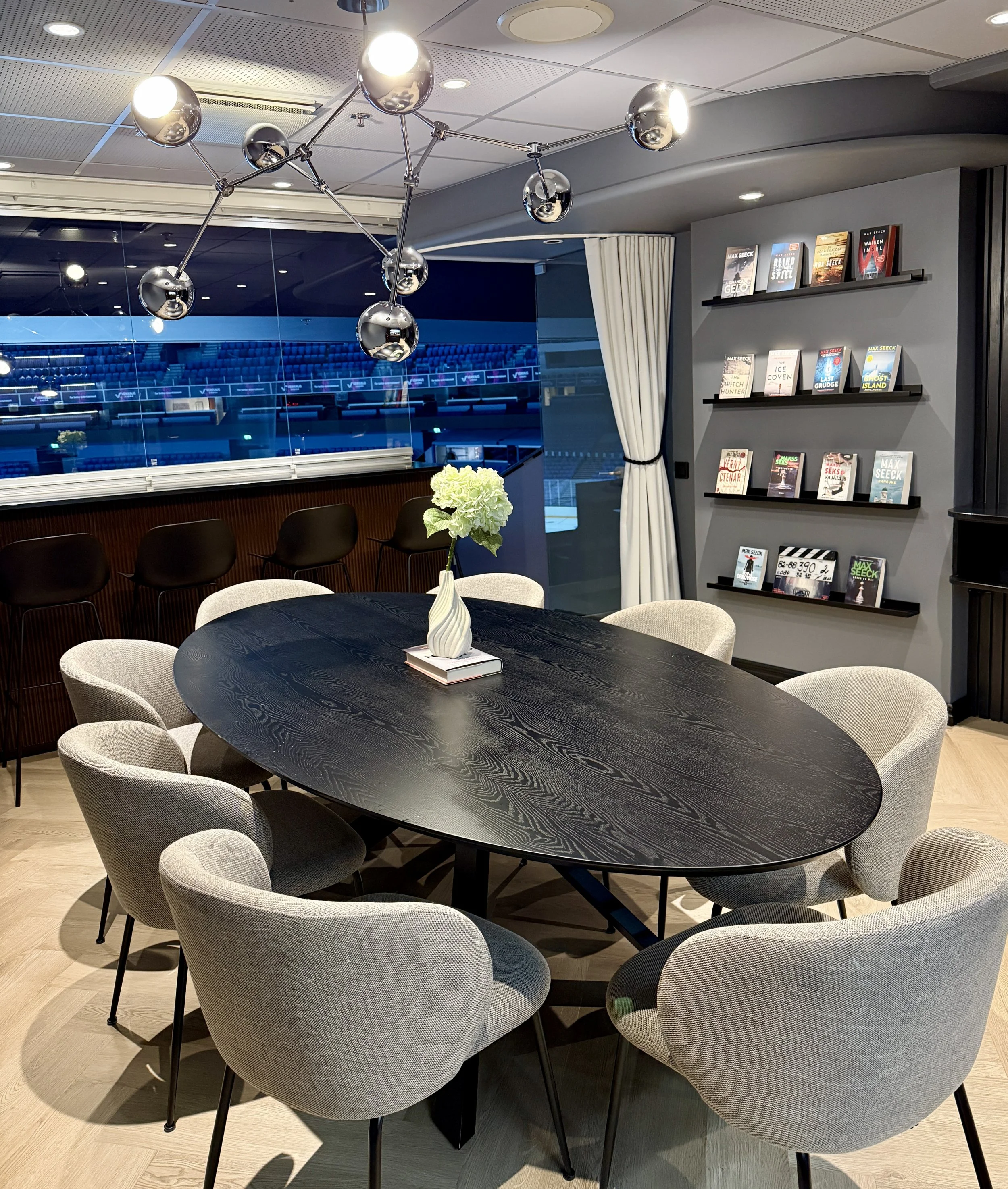 A modern conference room with a large black oval table surrounded by nine light gray upholstered chairs. A white vase with green hydrangeas and a book is on the table. There is a decorative chandelier with shiny metallic globes hanging from the ceili