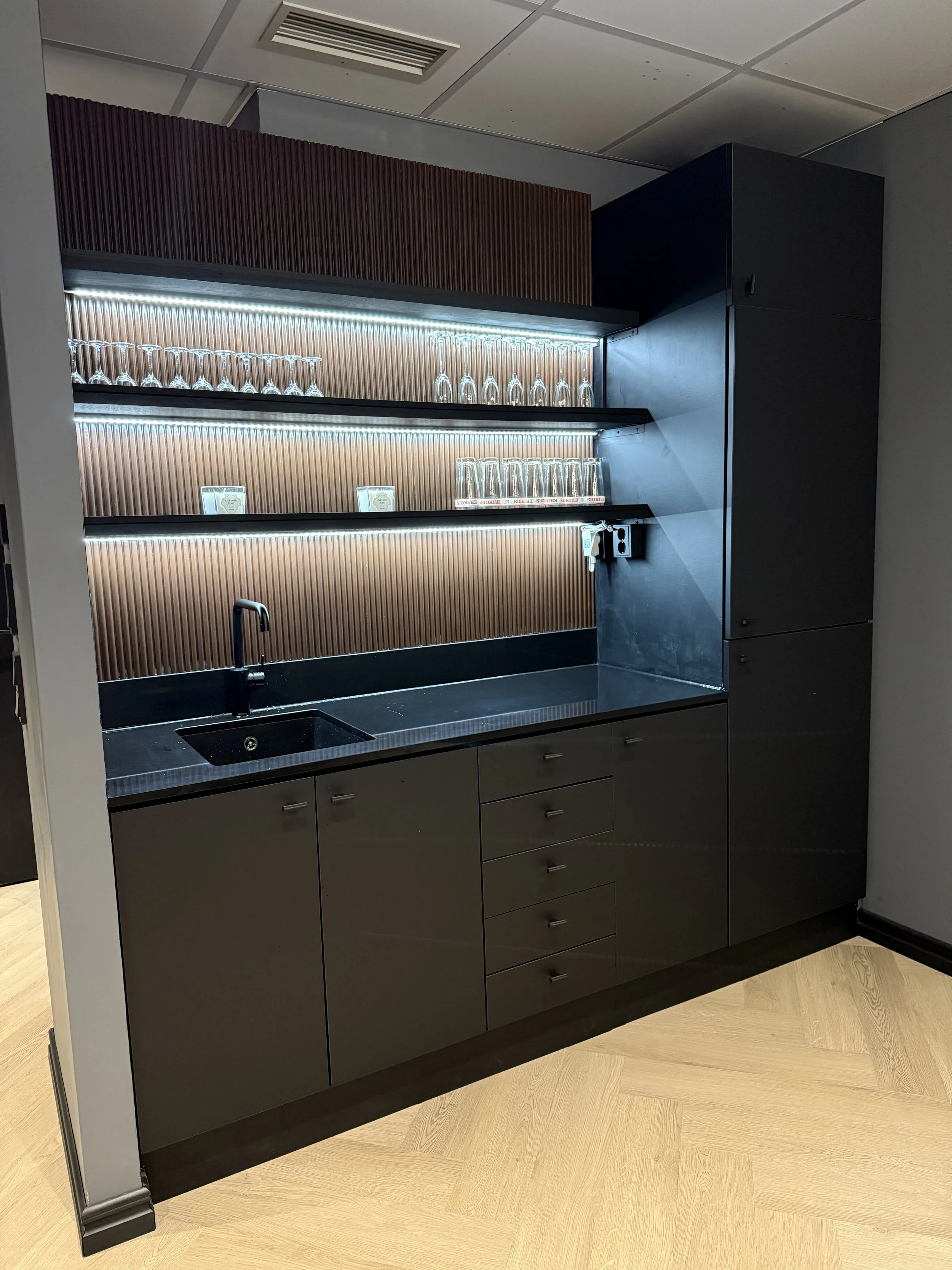 Modern kitchenette with dark cabinetry, a black countertop, a small sink, and shelves with glassware, illuminated with built-in lighting.
