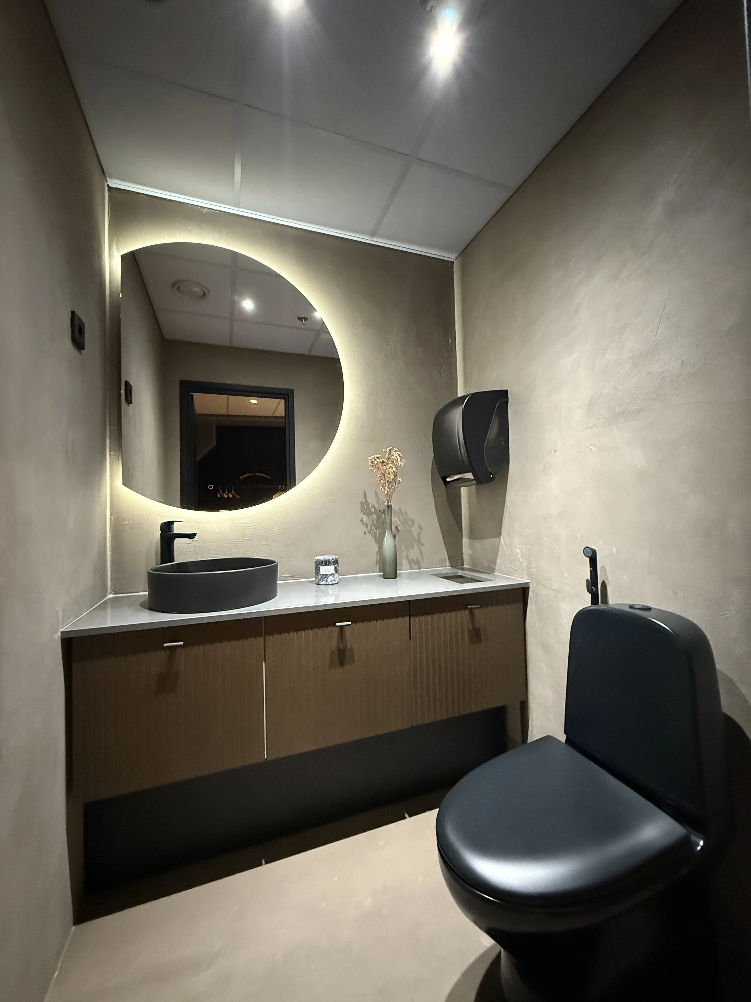 Modern bathroom with black sink, large round backlit mirror, black toilet, decorative vase with flowers, and a hand dryer on the wall.