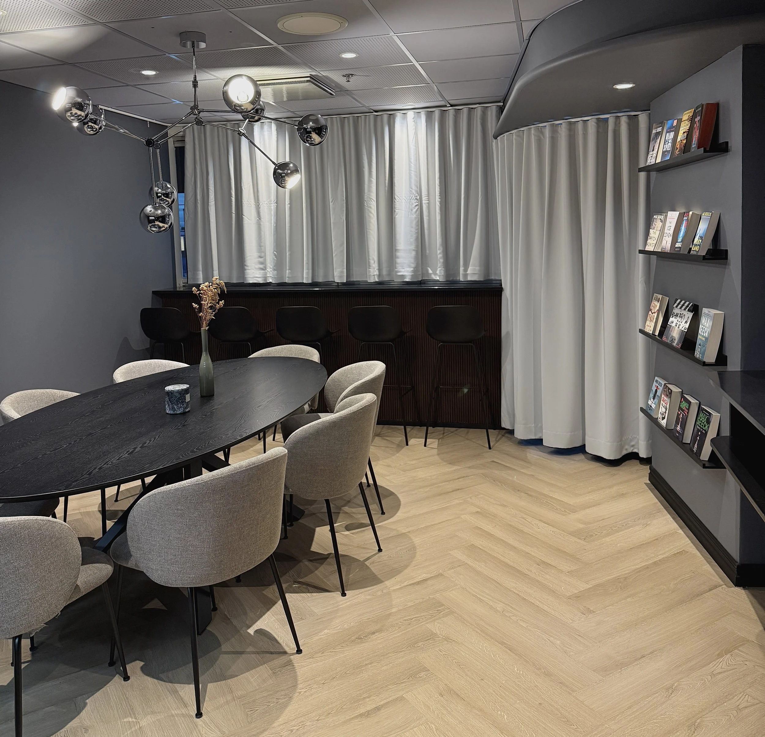 A modern lounge area with a black oval table surrounded by beige upholstered chairs, a decorative vase with dried flowers, a dark wood bar with black bar stools, a bookshelf filled with books, and contemporary ceiling lighting fixtures in a cozy sett