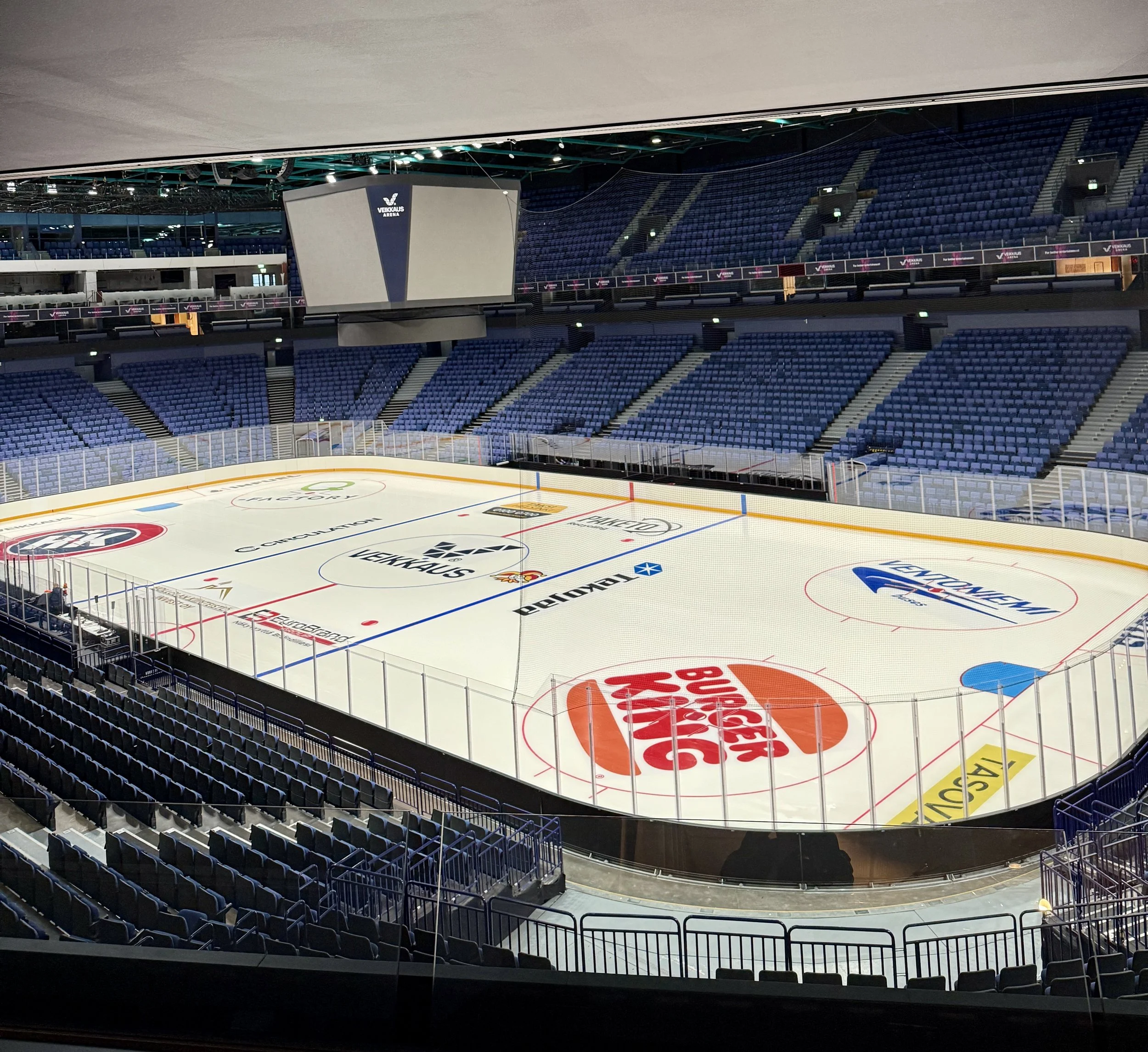 Empty ice hockey arena with blue seats, an ice rink with sponsor logos, and a large scoreboard hanging above.