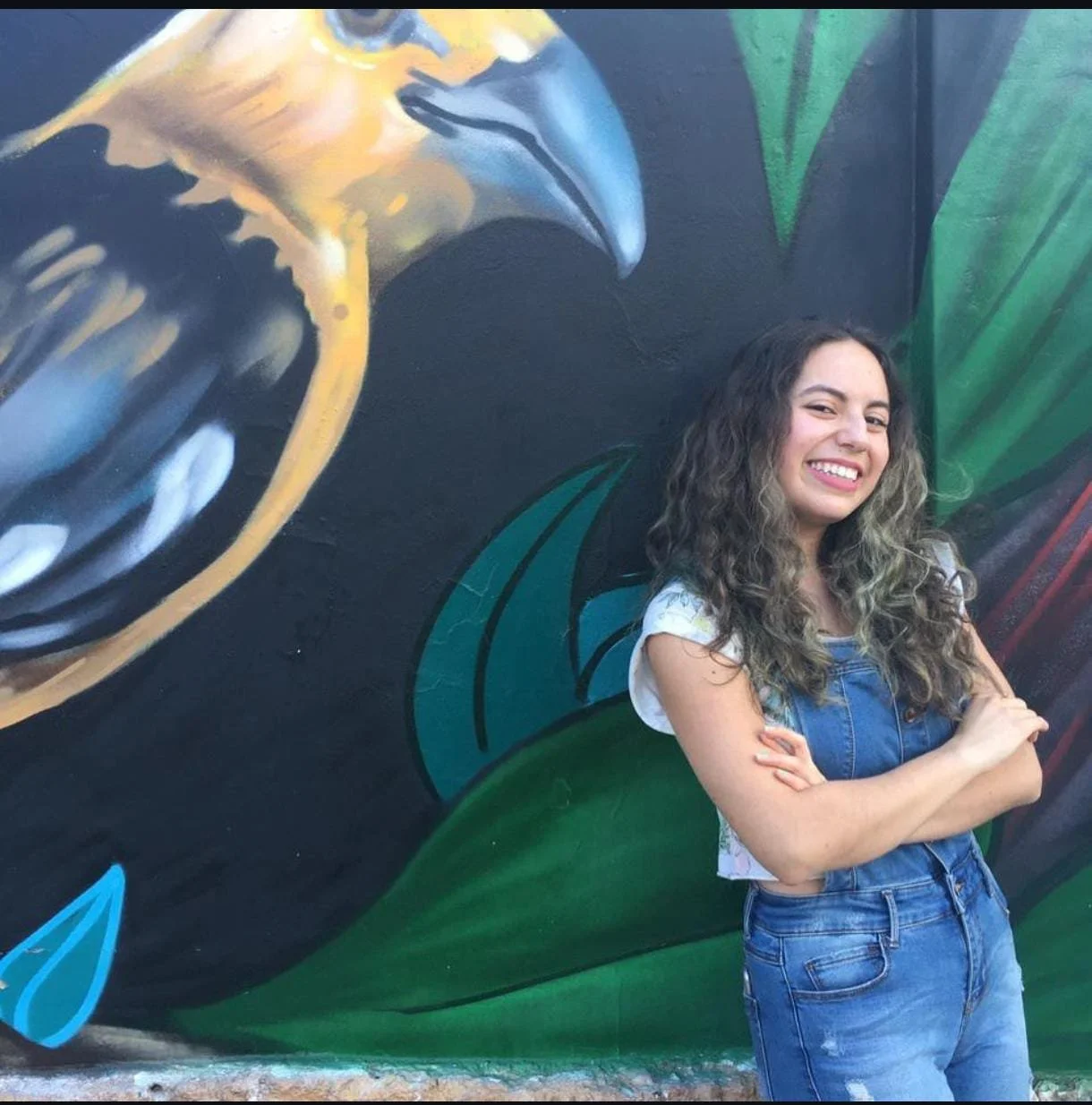 Junge Frau mit lockigem Haar posiert vor einer bunten Wandmalerei, die einen Vogel und Blätter zeigt.