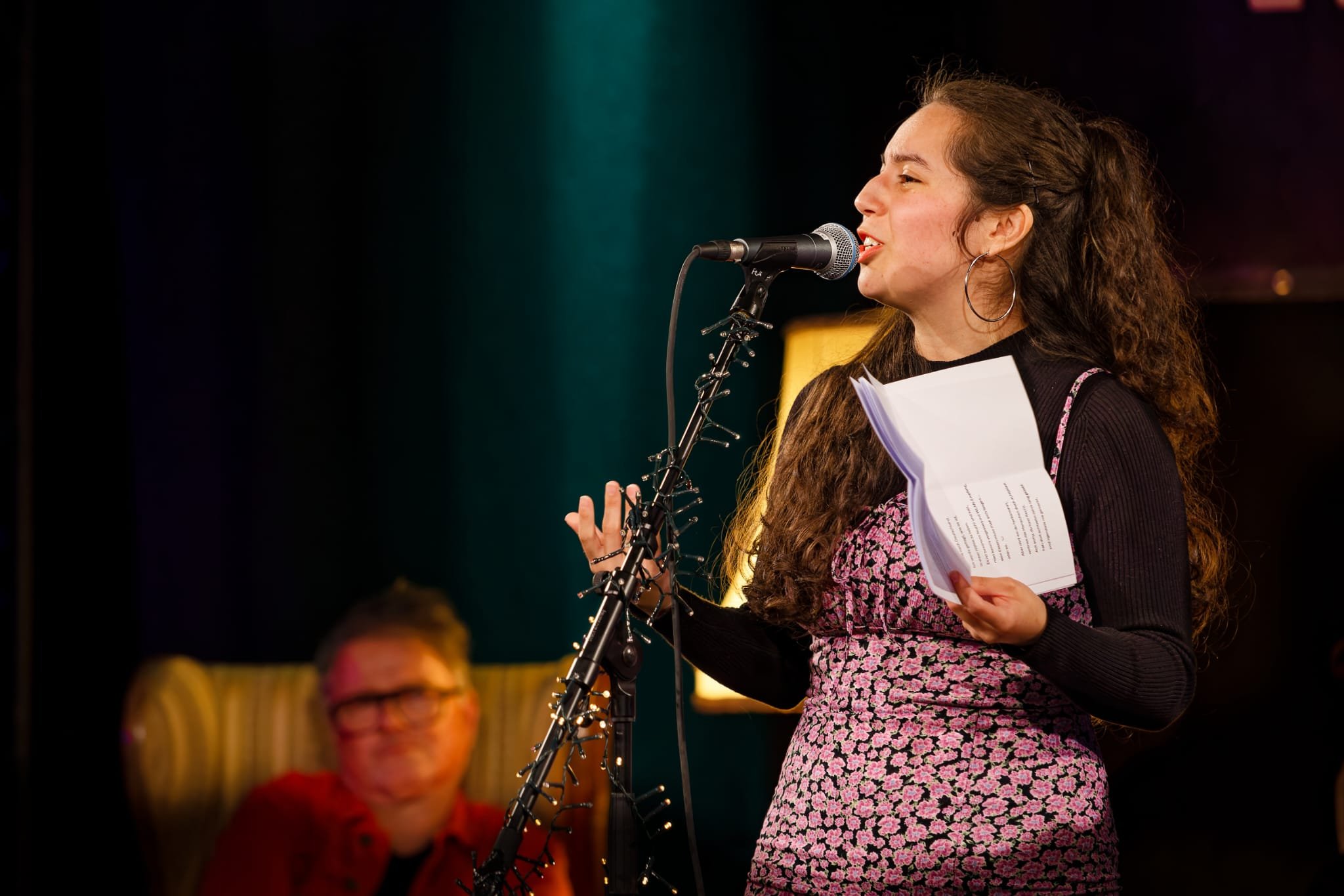 Junge Frau mit langen, lockigen Haaren, trägt einen Text bei einem Poetry Slam auf der Bühne vor.