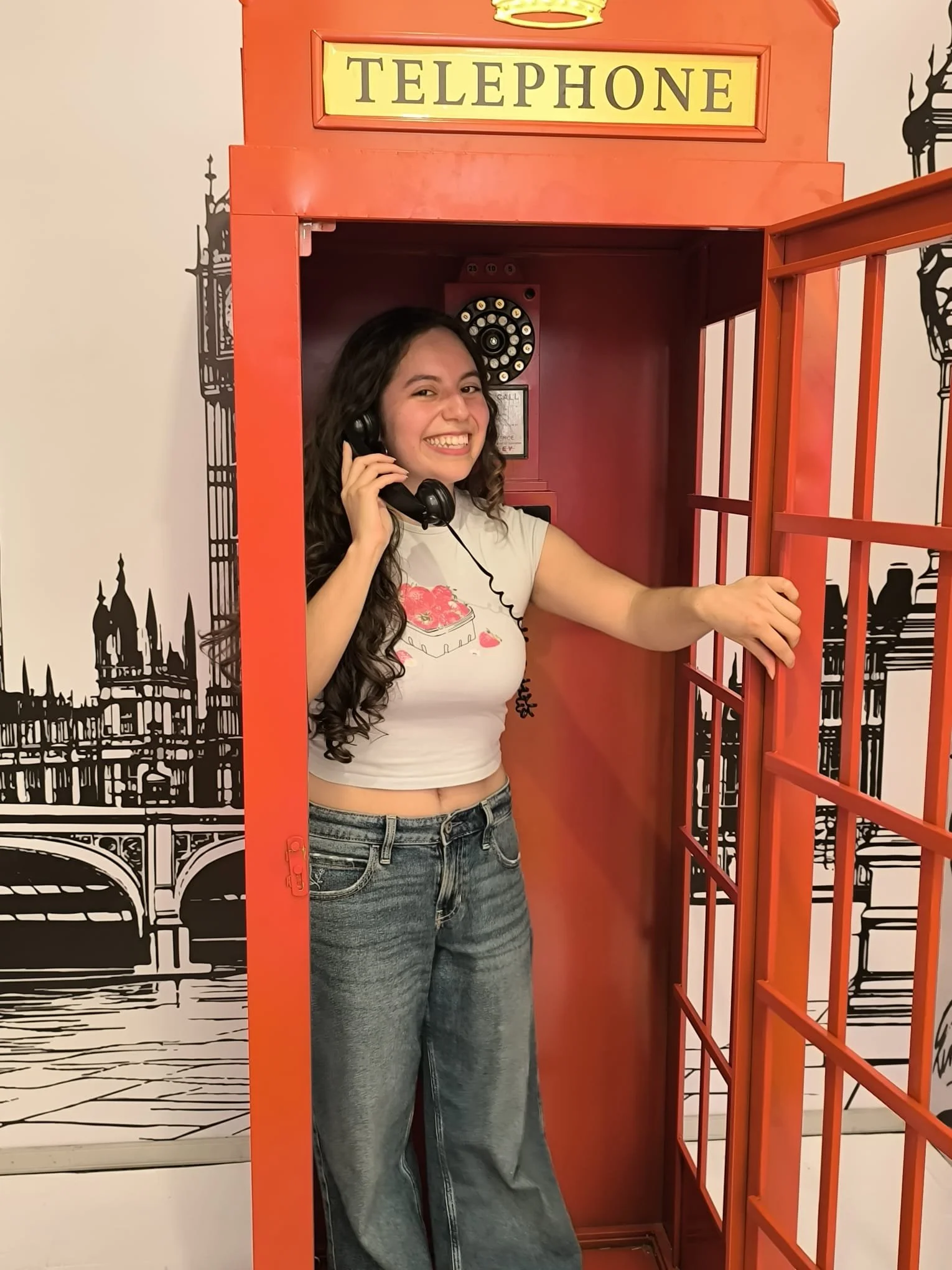 Junge Frau steht in einer roten Telefonzelle, spricht ins Telefon und lächelt.