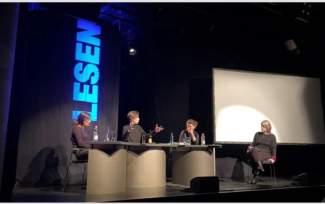 Vier Menschen sitzen während einer Diskussion auf einer Bühne, es gibt einen Tisch mit Wasserflaschen, im Hintergrund eine große weiße Leinwand und eine vertikale blaue Leuchttafel.