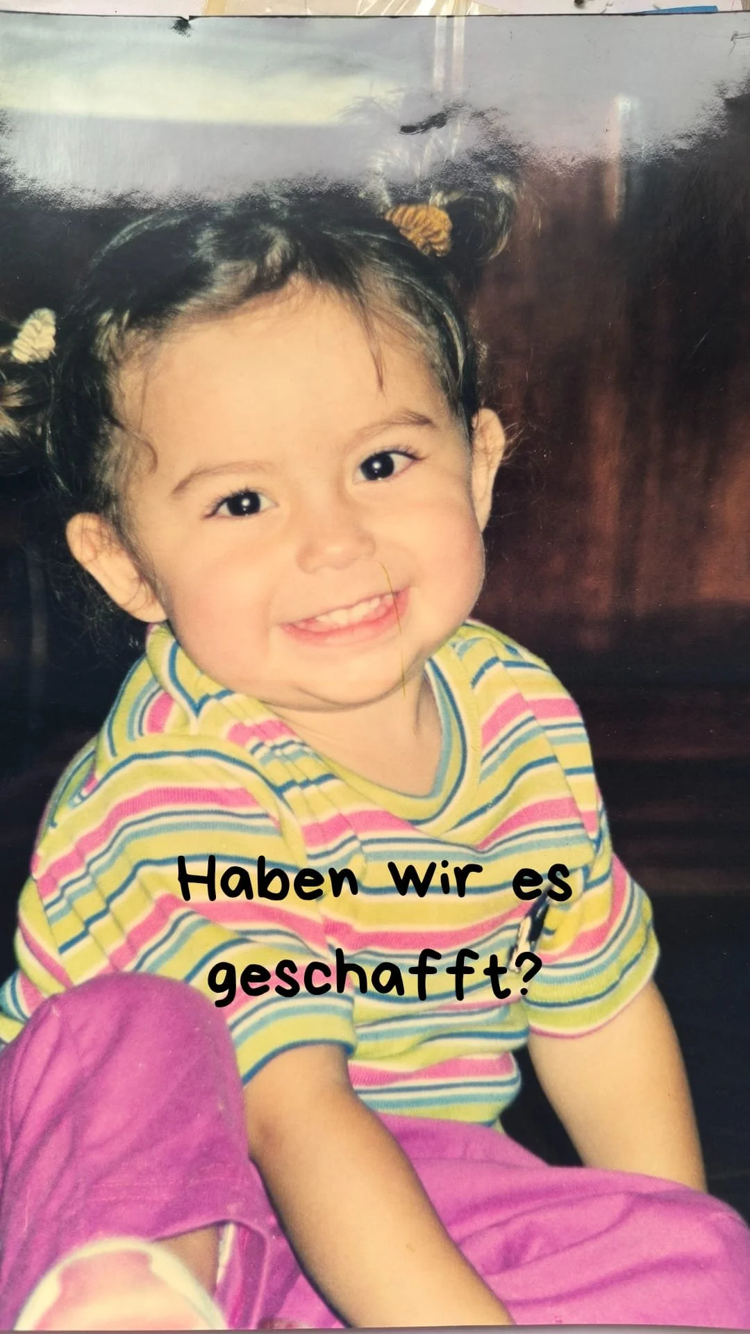 Ein lachendes kleines Mädchen mit Zöpfen, in buntem gestreiften T-Shirt und pinken Hosen, sitzt vor einem dunklen Hintergrund.