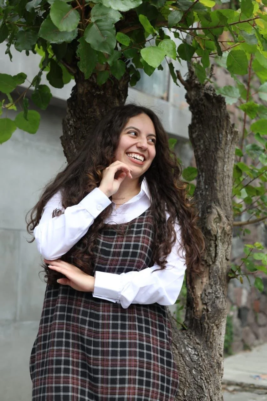 Junge Frau mit langem, lockigem Haar, lachend, steht neben einem Baum, trägt weiße Bluse und kariertes Kleid, im Freien.