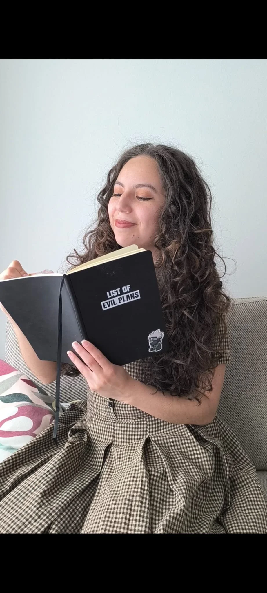 Junge Frau mit lockigem Haar schreibt ein Gedicht auf einem Heft. Sie lächelt und sitzt auf einem Sofa.