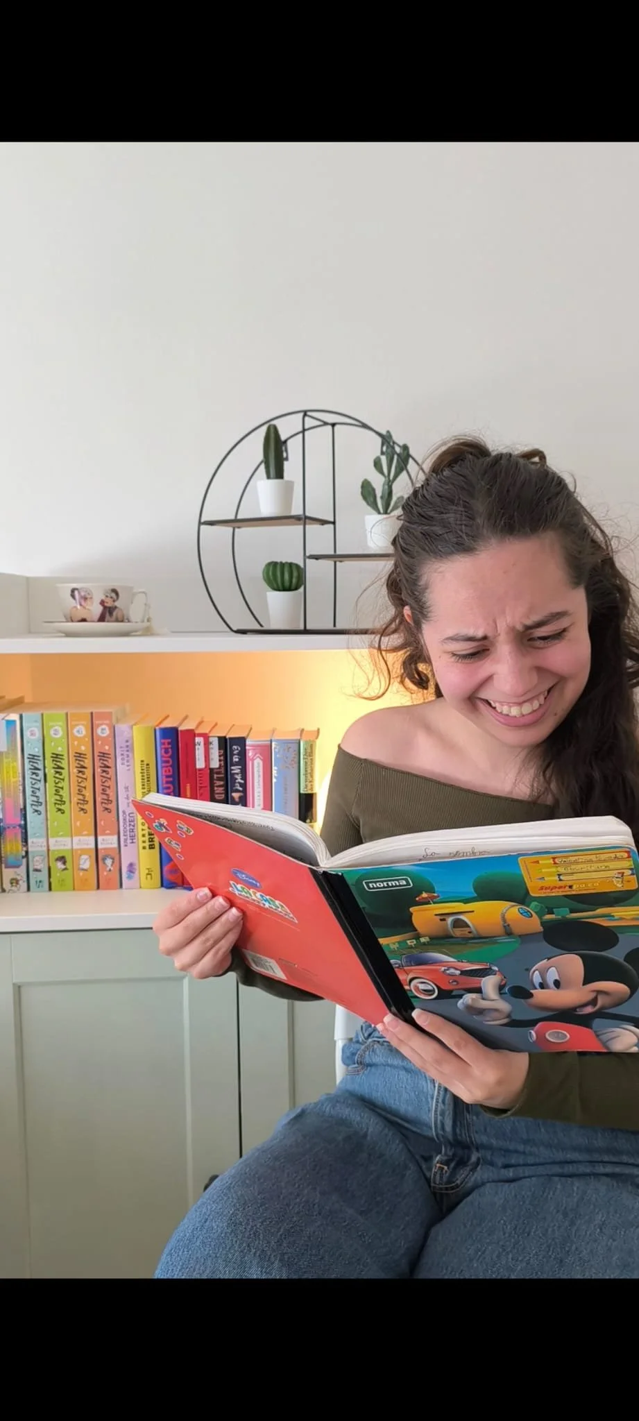 Eine junge Frau sitzt in einem Raum und liest aus einem Heft vor. Sie sieht amüsiert und überrascht aus.