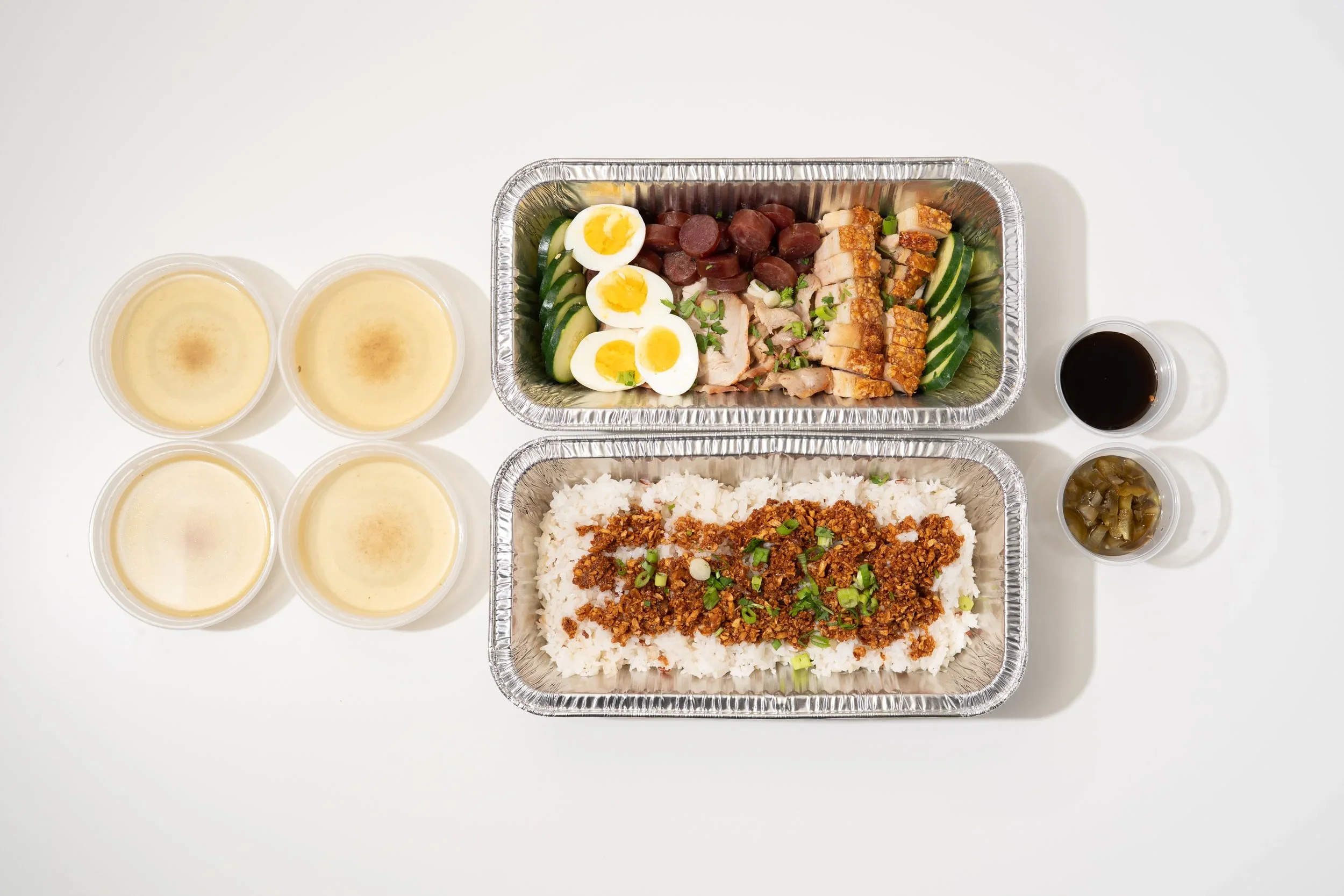 Meal in aluminum containers with rice topped with seasoned ground meat, greens, and green onion; side of cucumber, sliced chicken, and sausage with garnishes; small cups of soy sauce, sweet sauce, and pickled vegetables.