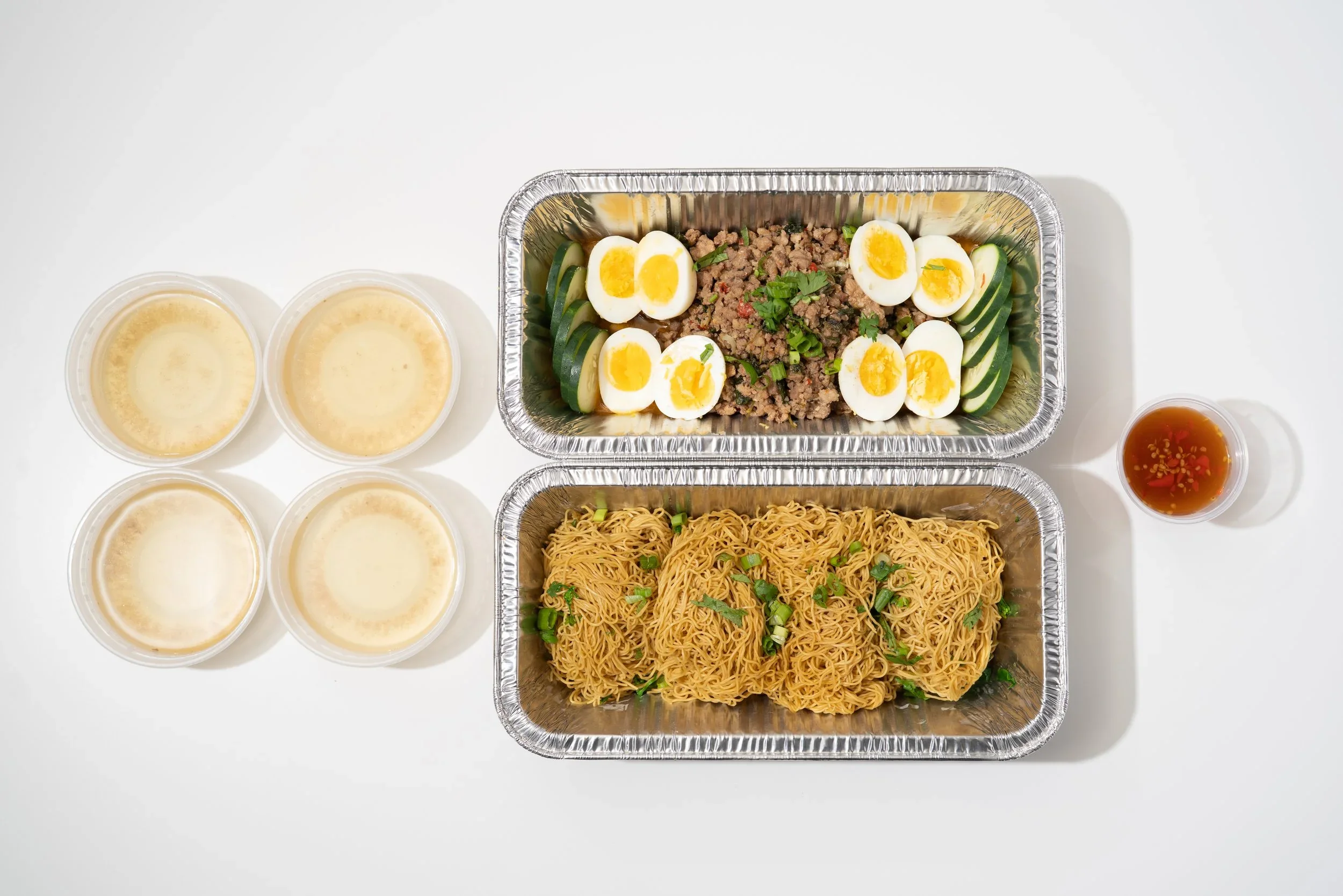 Two aluminum trays of Asian food, one with cooked noodles and the other with chopped meat, sliced cucumbers, and halved boiled eggs, surrounded by four small bowls of sauce or broth and a small cup of red chili sauce.