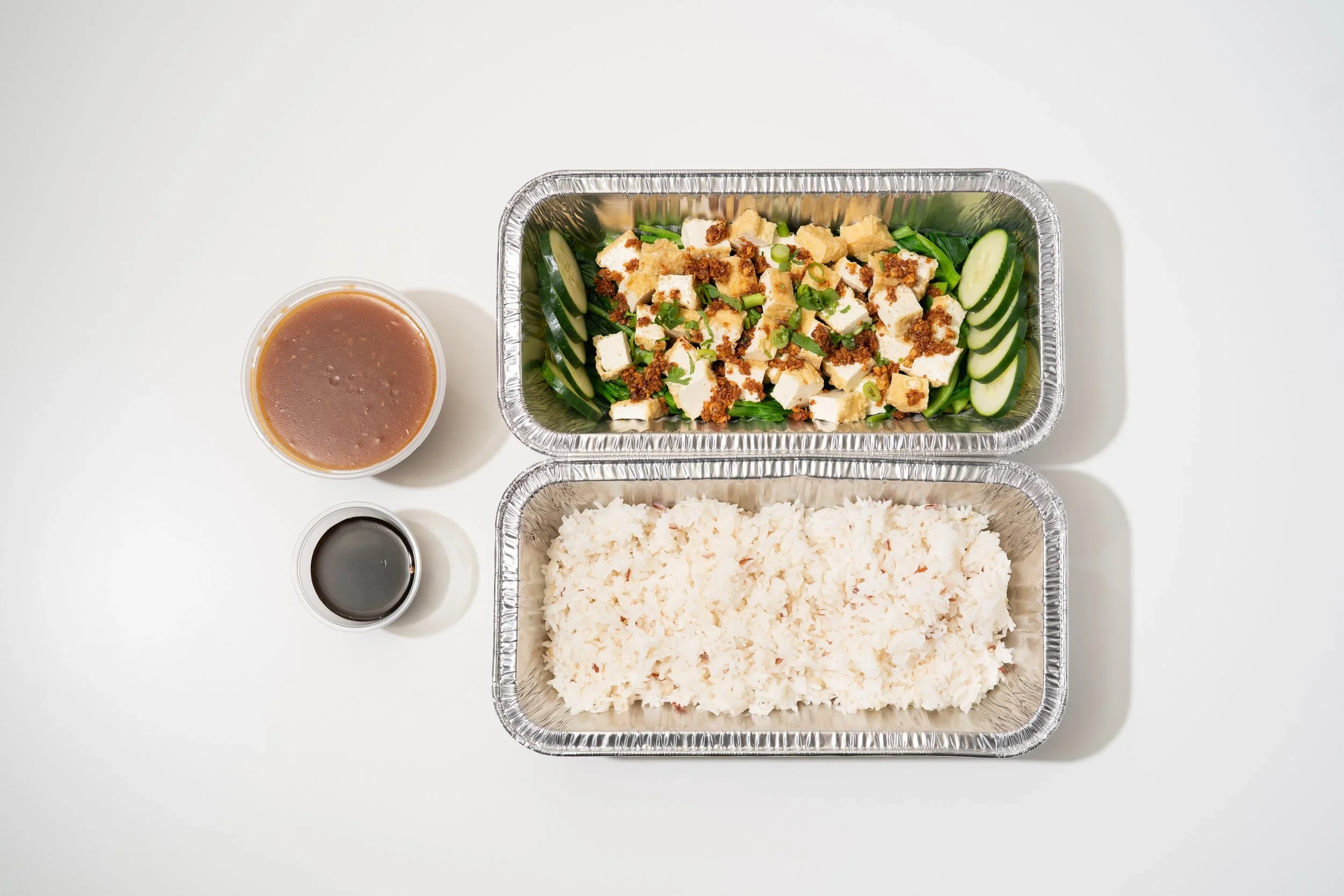 Meal with white rice, tofu salad with cucumbers and green onions, two small containers of soy sauce and dressing on a white surface.