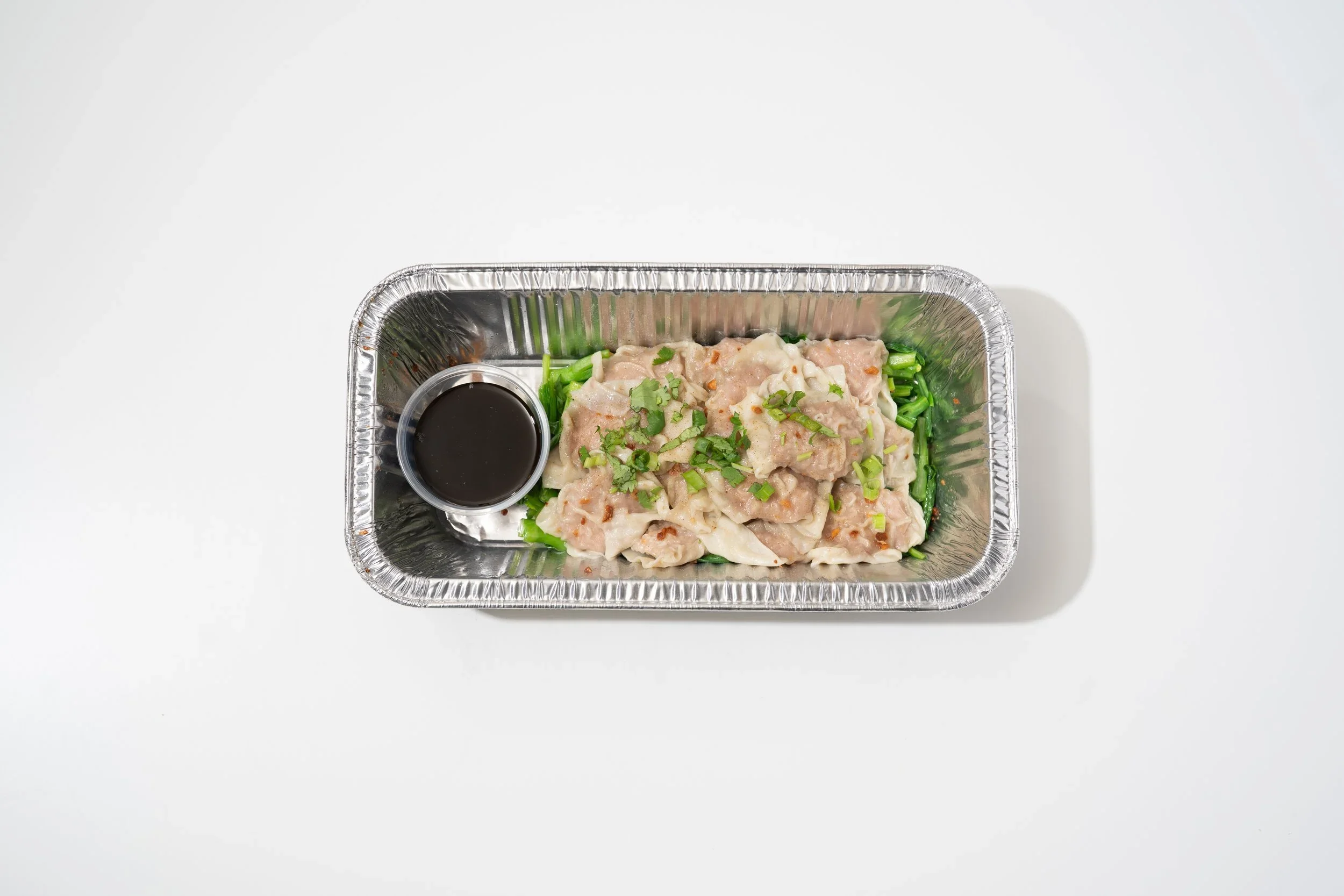 A rectangular aluminum takeout container with chopped green vegetables and a portion of chicken or fish topped with chopped green onions, accompanied by a small cup of soy sauce on a white surface.