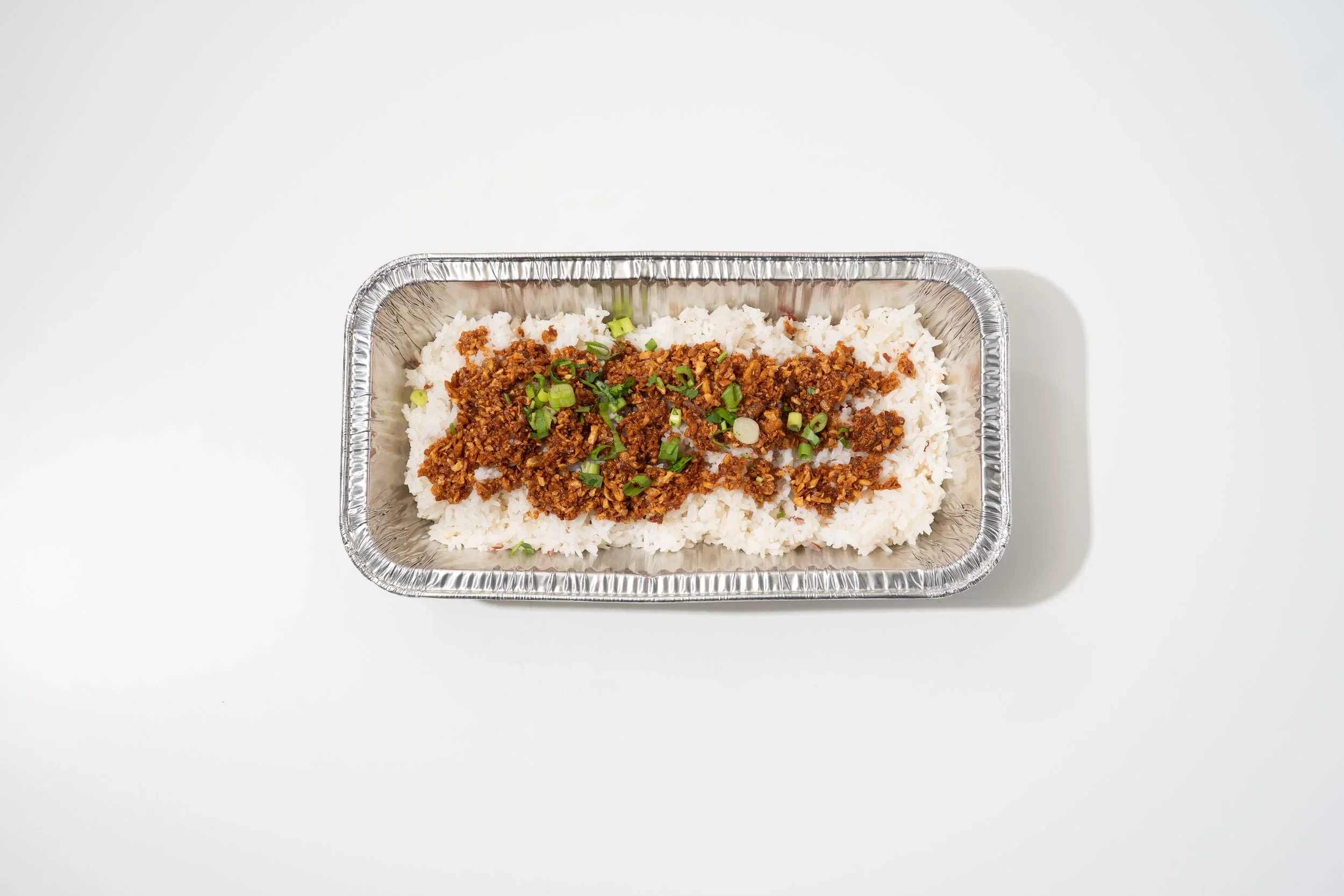 Foil container of white rice topped with ground meat and chopped green onions, on a white surface.