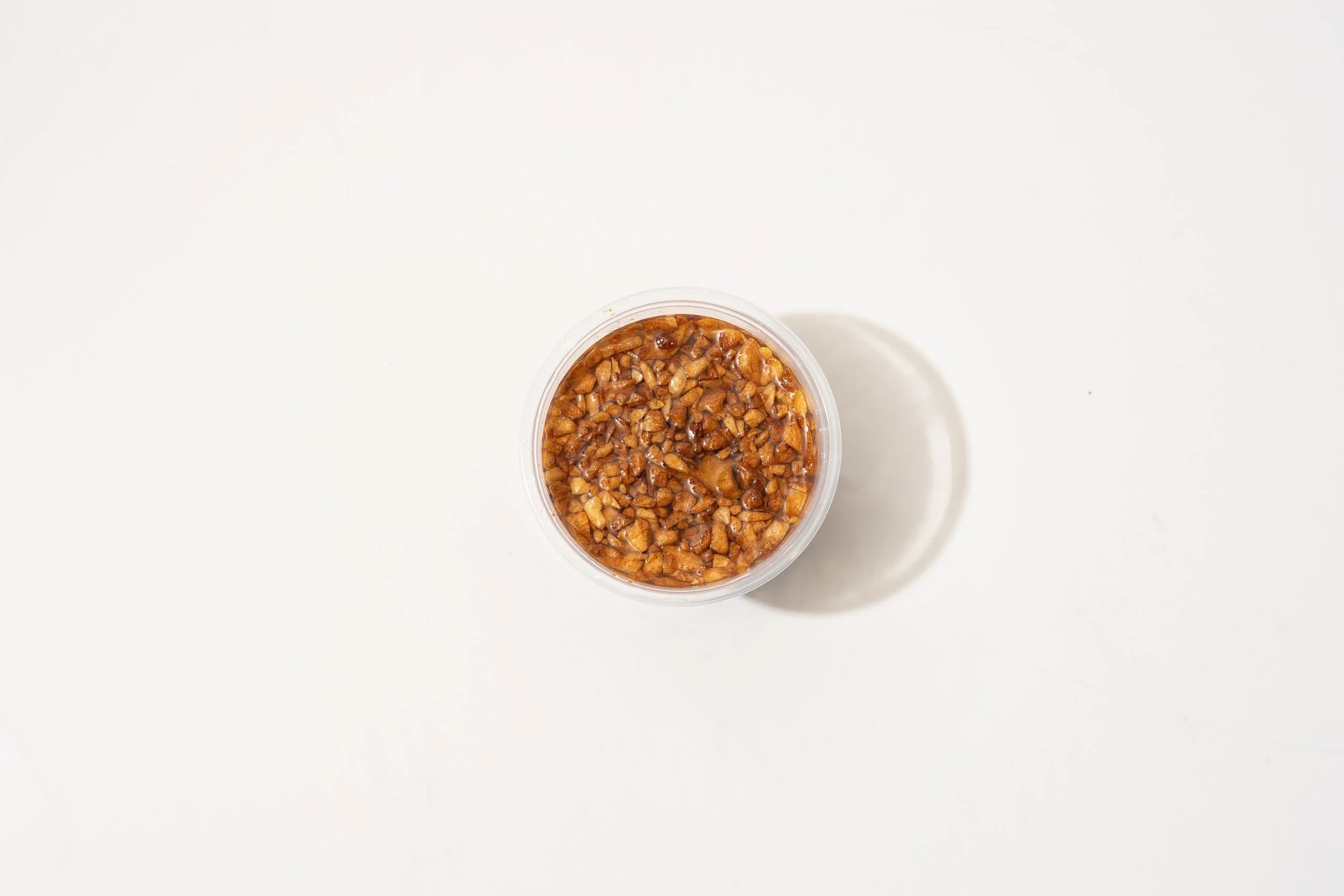 Top view of a small container filled with chopped nuts in syrup on a white surface, casting a shadow.