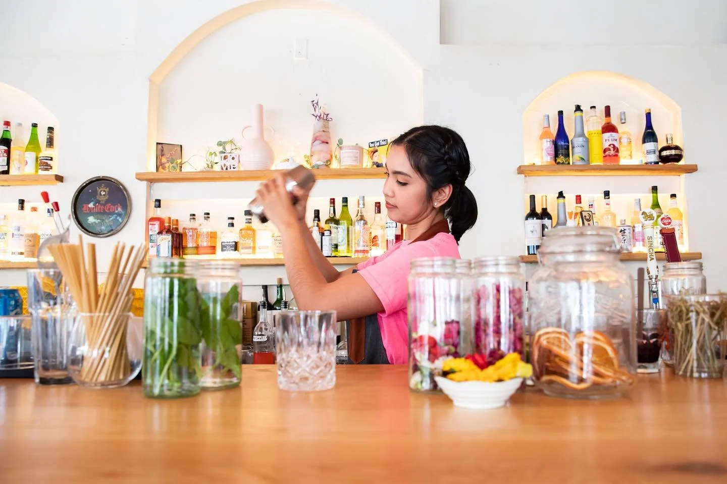 A young woman behind a bar counter with various jars containing colorful dried fruit and herbs, arranging bottles on shelves with liquor bottles and decorative items in the background.