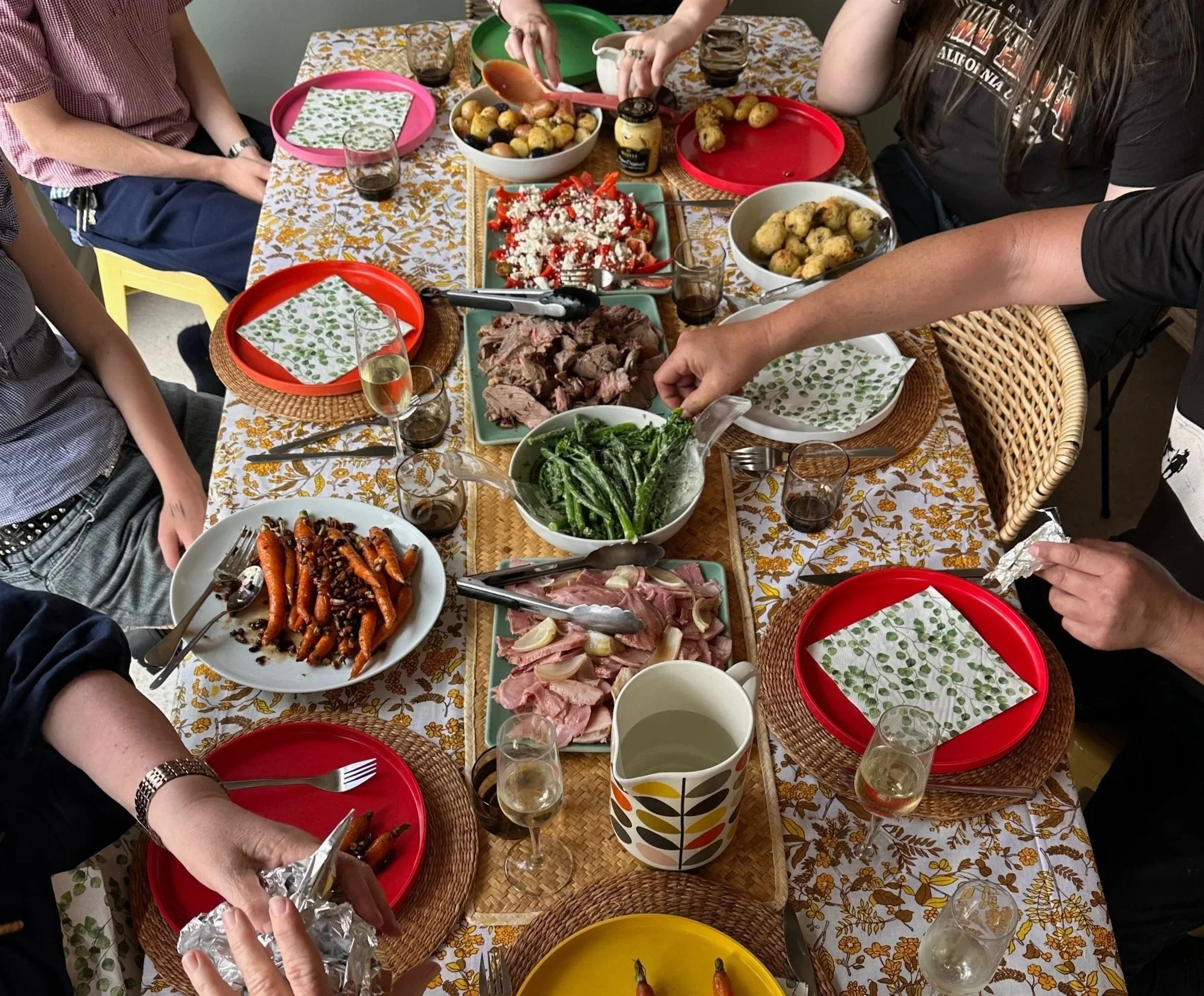 A family or friends gathered around a table with various dishes for a meal, including vegetables, meat, and side dishes, with plates, glasses, and utensils on a tablecloth.