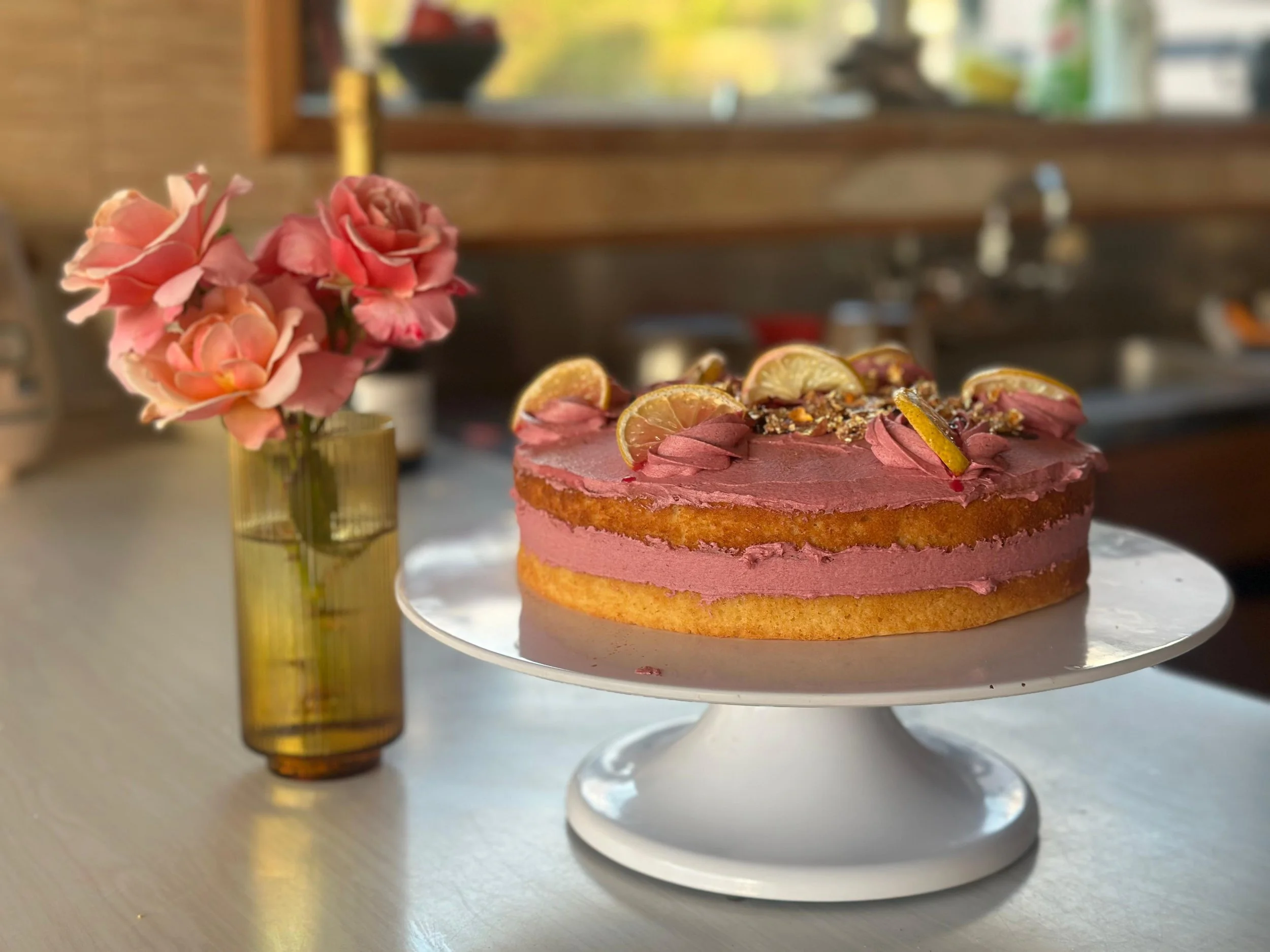 A layered cake with pink frosting, decorated with lemon slices and nuts, placed on a white cake stand next to a pink flower in a yellow glass vase.