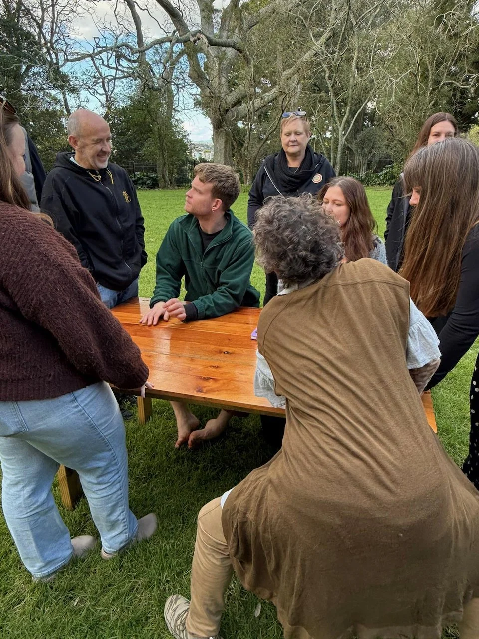 A group of people gathered outdoors around a wooden table, engaged in conversation, with a man sitting at the table and others standing around him, in a park-like setting with trees and grass.