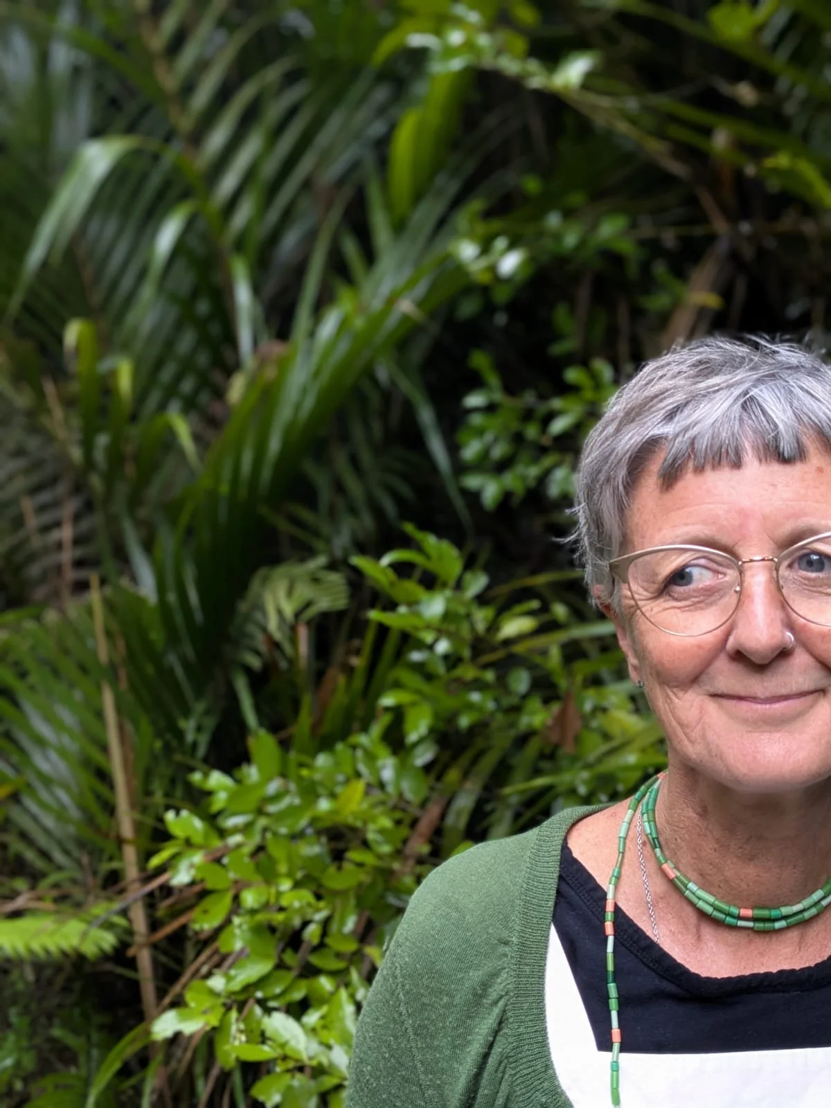 A woman with short gray hair, glasses, and a nose ring smiling outdoors in front of lush green tropical plants. She is wearing a green cardigan and beaded necklaces.
