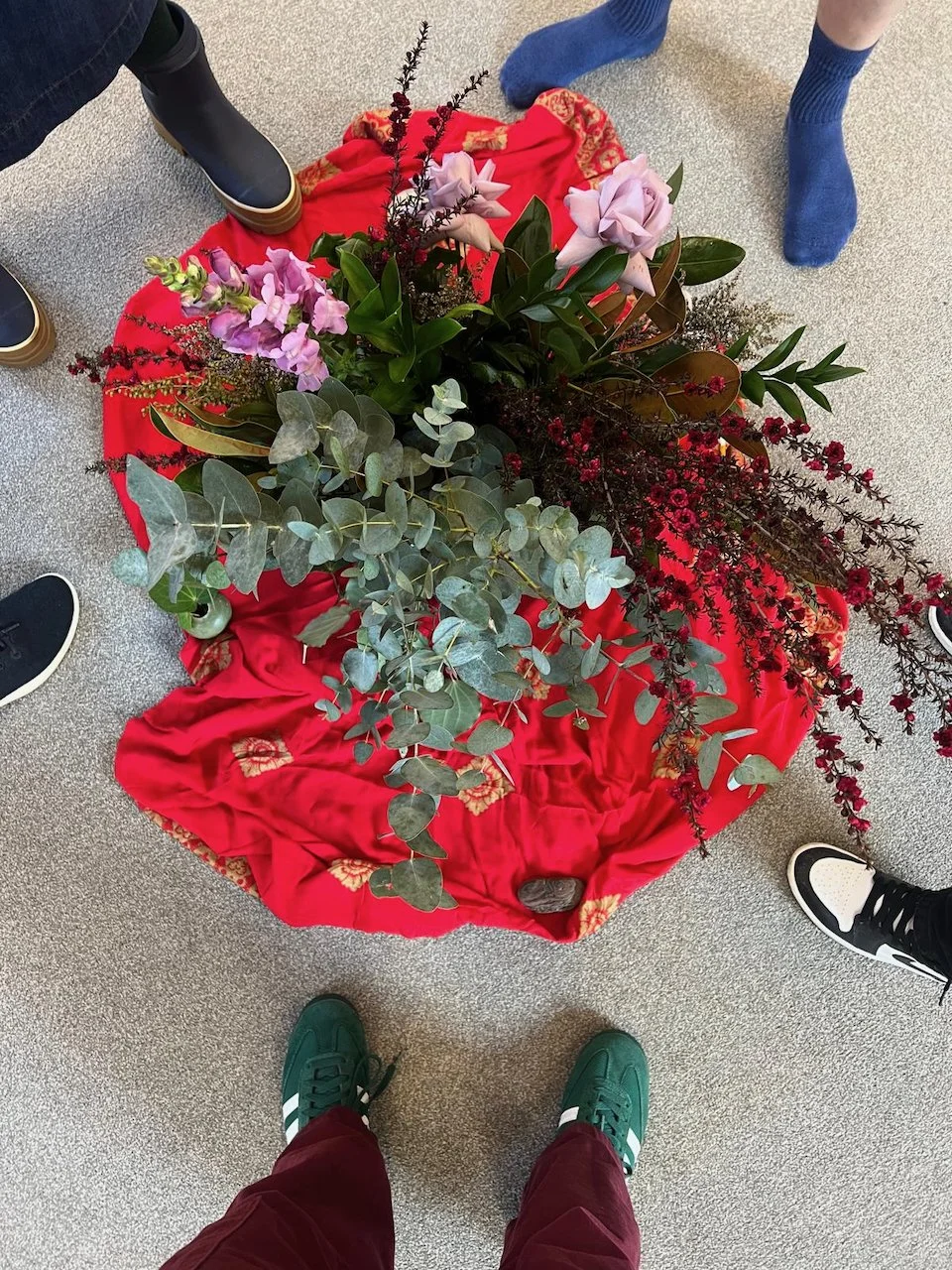 People standing around a large floral arrangement on a red cloth on the floor, containing pink roses, purple snapdragons, eucalyptus leaves, and other greenery.