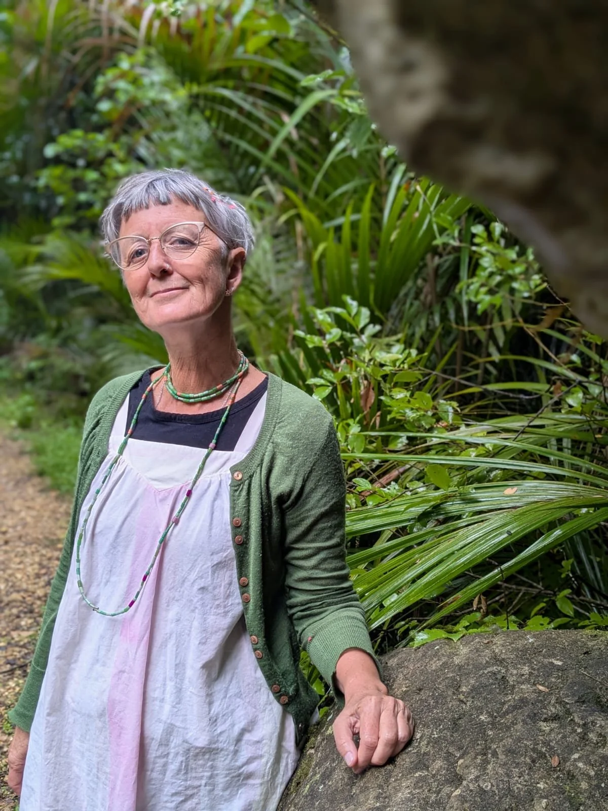 An older woman with short gray hair, glasses, and a slight smile, standing outdoors in a lush, green, tropical garden, resting her hand on a large rock.