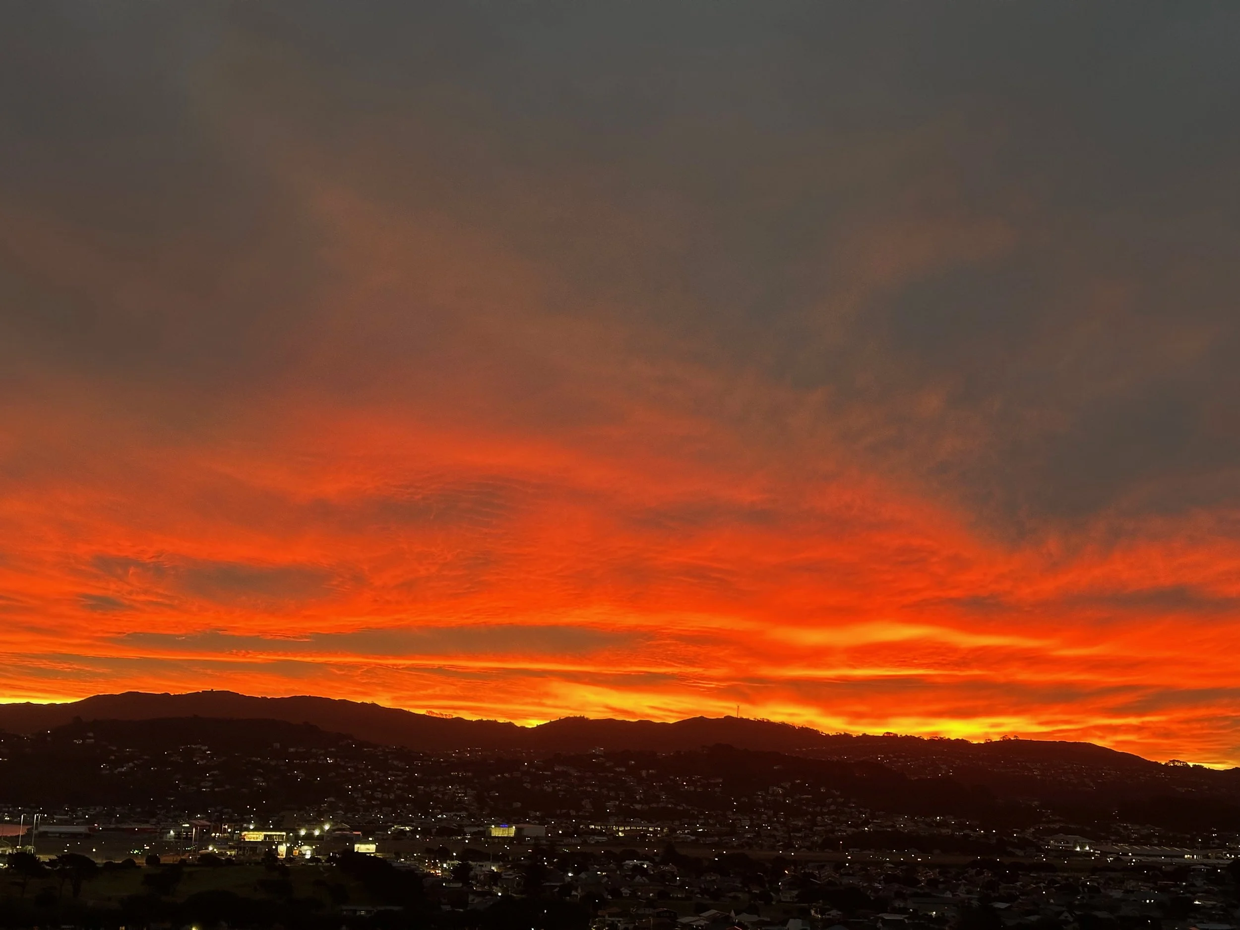 A vibrant sunset over a hilly cityscape, with orange and red hues illuminating the sky and silhouetted hills and buildings in the foreground.