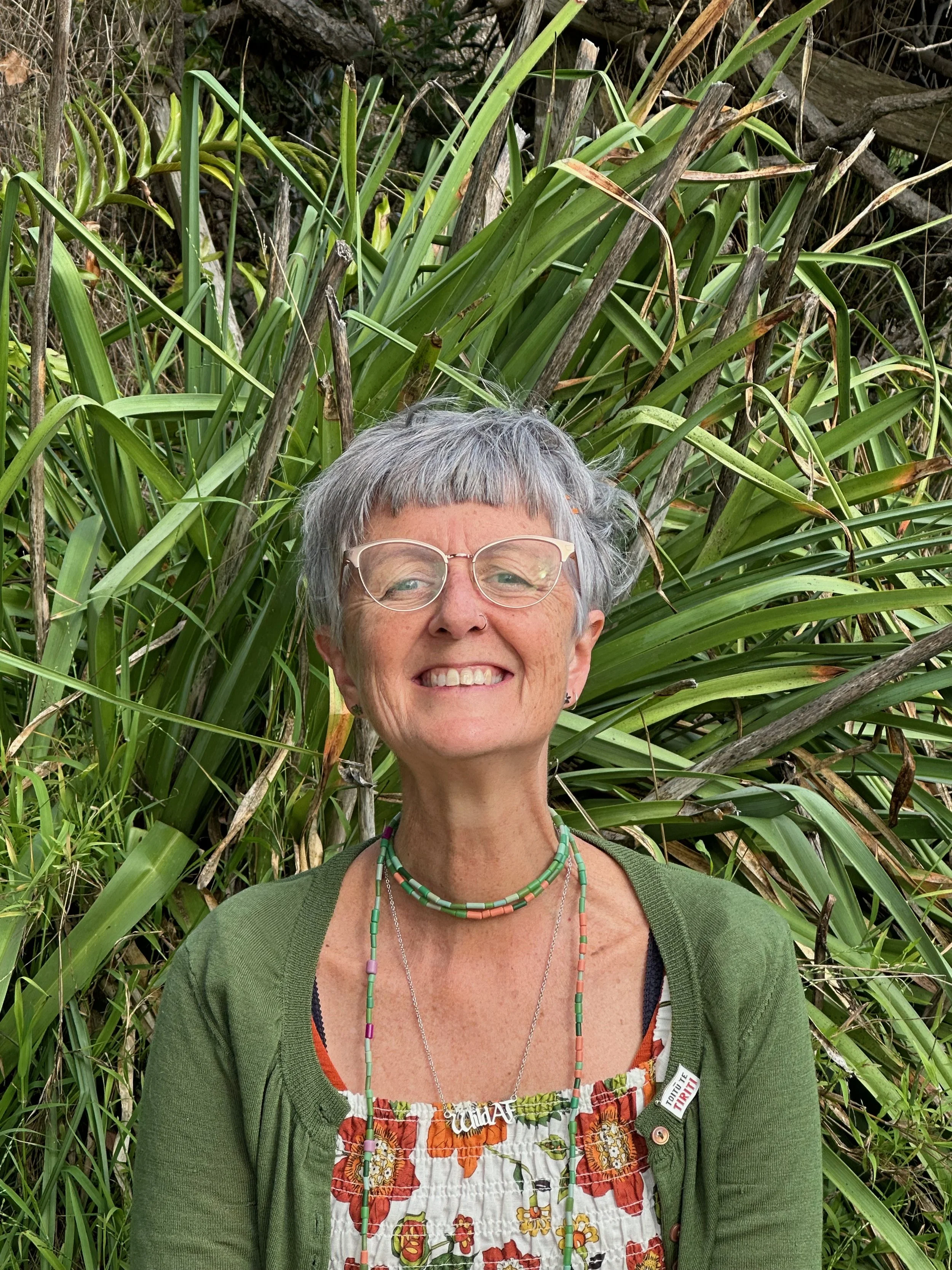 Smiling woman with gray hair and glasses standing in front of tall green plants, wearing a colorful dress, green cardigan, and colorful beaded necklaces.