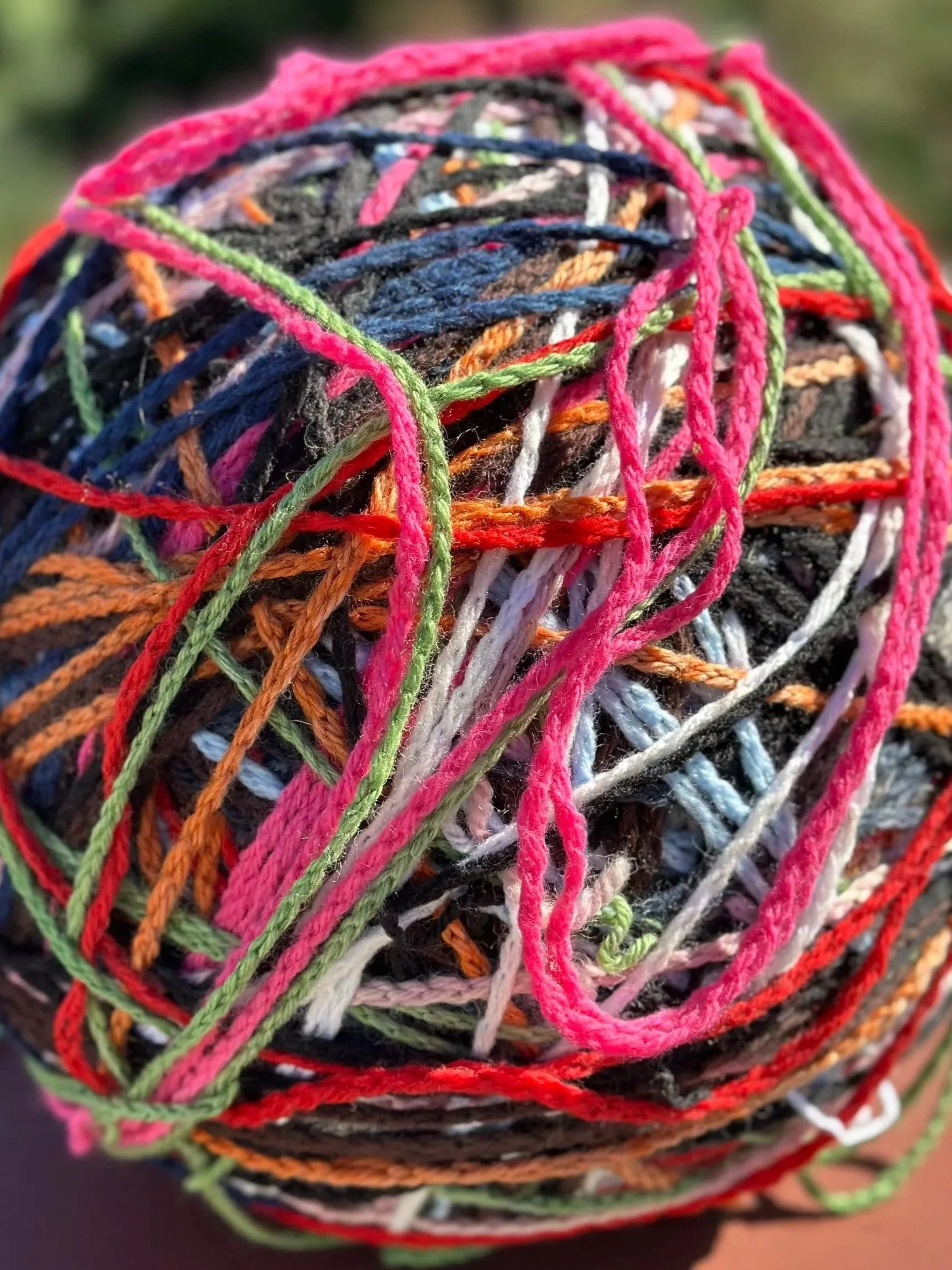 Close-up of a tangled, colorful ball of yarn with various strands in pink, green, orange, white, gray, black, and blue.