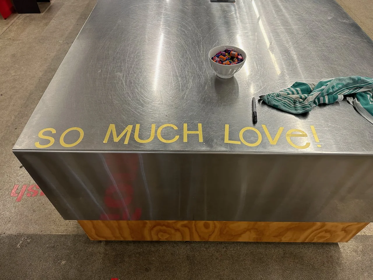 A stainless steel table with yellow text on the front that says, 'SO MUCH LOVE!'. On the table, there is a bowl of assorted candies, a black marker, and a crumpled striped cloth. The background includes a concrete floor with pink writing on it.