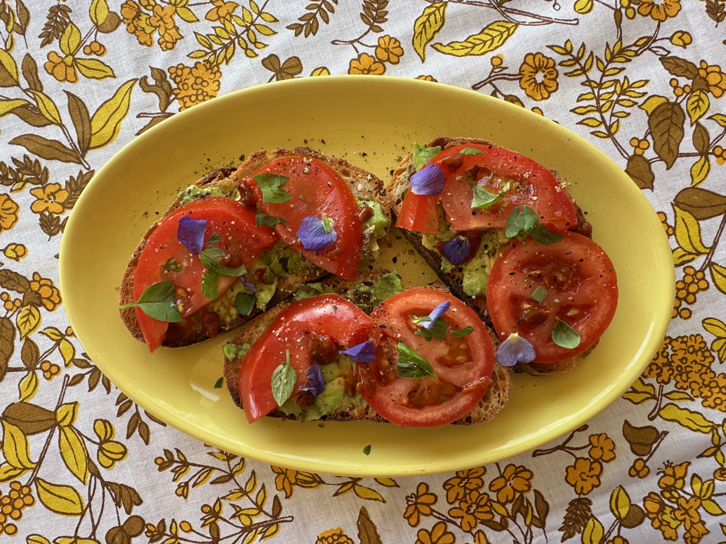 Open-faced avocado toast topped with sliced tomatoes, fresh herbs, and edible flower petals on a yellow plate.