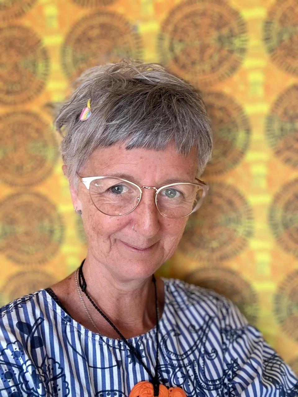 A smiling woman with short gray hair, glasses, and a striped blue and white top, standing in front of a colorful, patterned background.