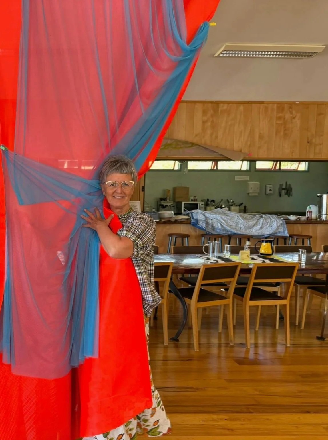 Middle-aged woman with glasses, short gray hair, wearing a checkered shirt and floral skirt, smiling and peeking out from behind a colorful, sheer fabric in a dining or meeting space with wooden floors and tables set with glassware and coffee pots.