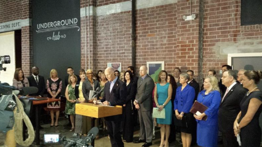 A group of people gathered in a brick-walled room, participating in a formal event or ceremony, with one man speaking at a wooden podium and others standing around him, some holding documents.