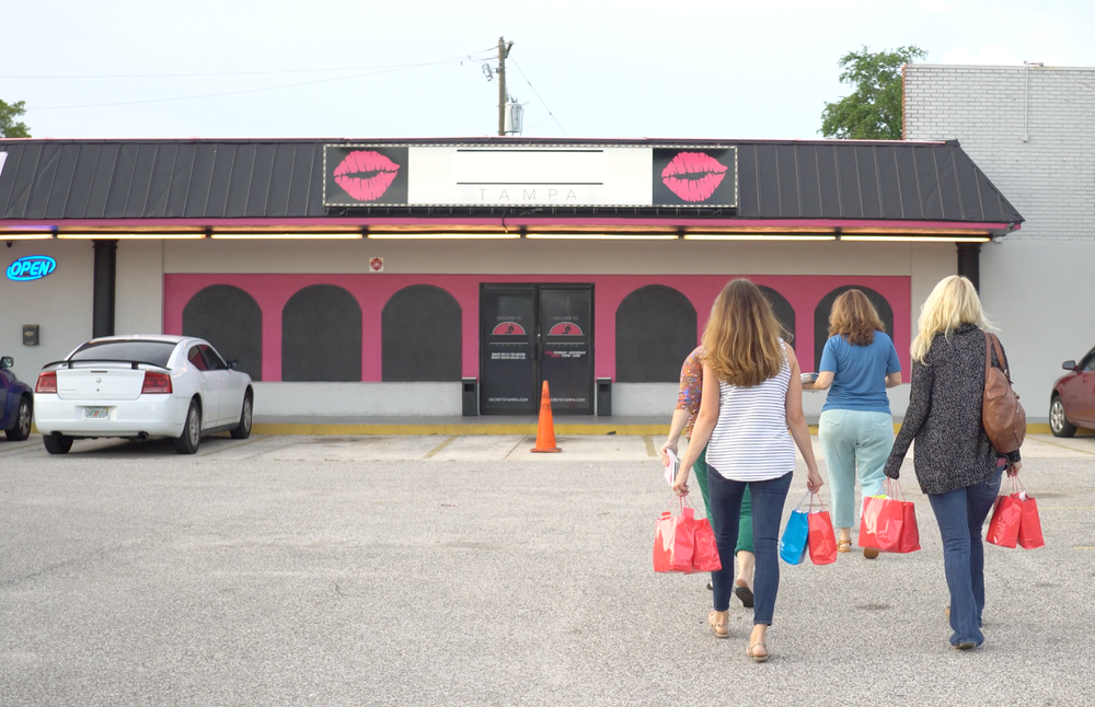 Four women walking towards a store with pink lipstick graphic signs, carrying shopping bags, in a parking lot.