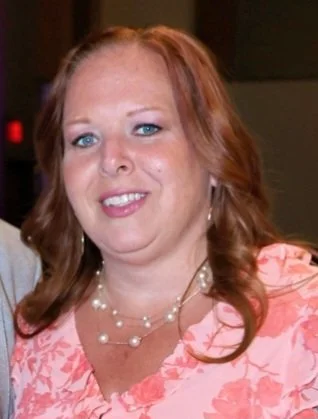 A woman with red hair, blue eyes, and fair skin, smiling, wearing a pink floral dress and layered pearl necklaces.
