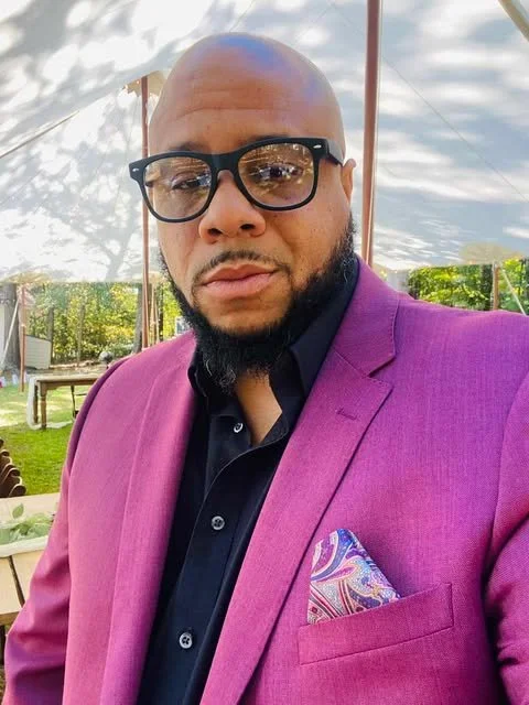 A man with a shaved head, beard, and glasses wearing a purple blazer over a black shirt and a colorful pocket square, standing outdoors under a white canopy.